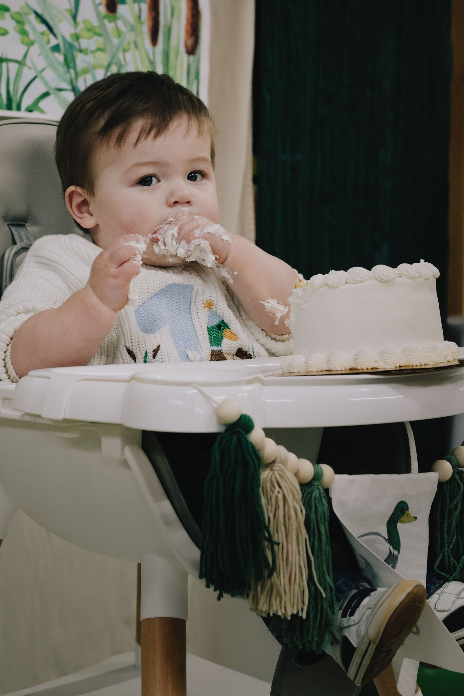 Baby in highchair plays messily with cake during what appears to be a first birthday celebration.