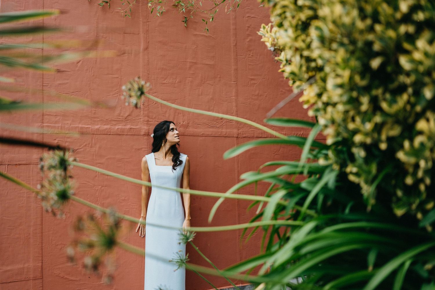 Bride in white dress posing against terracotta wall with green foliage.
