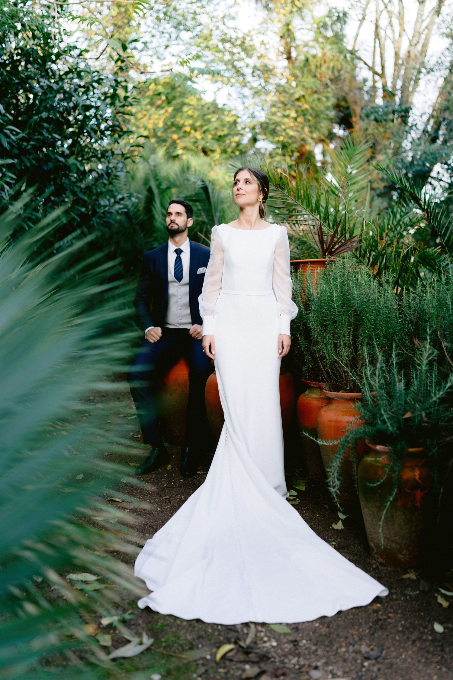 Wedding couple stands in lush garden setting surrounded by potted plants and greenery.