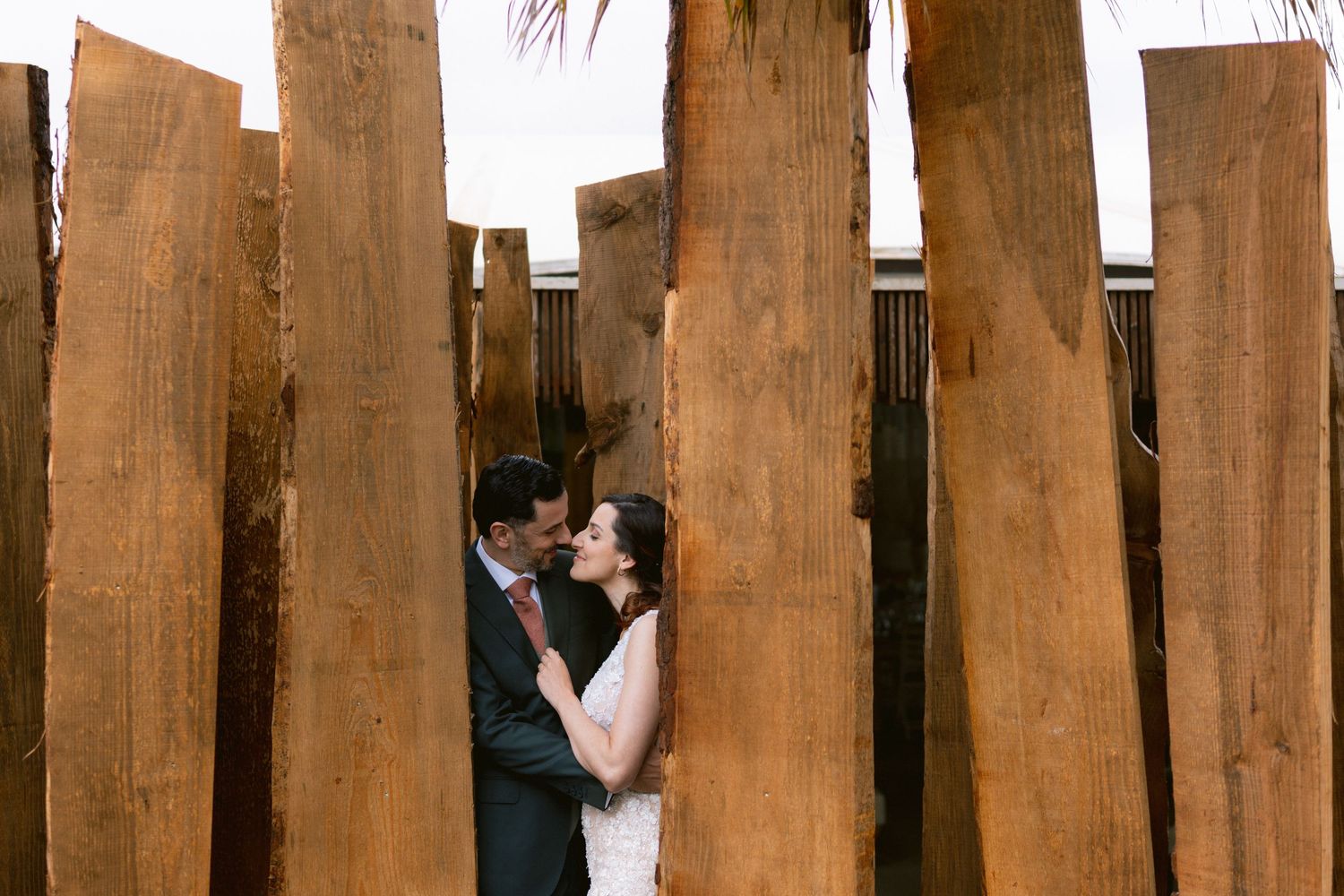 A couple embraces between rustic wooden fence posts in natural lighting.