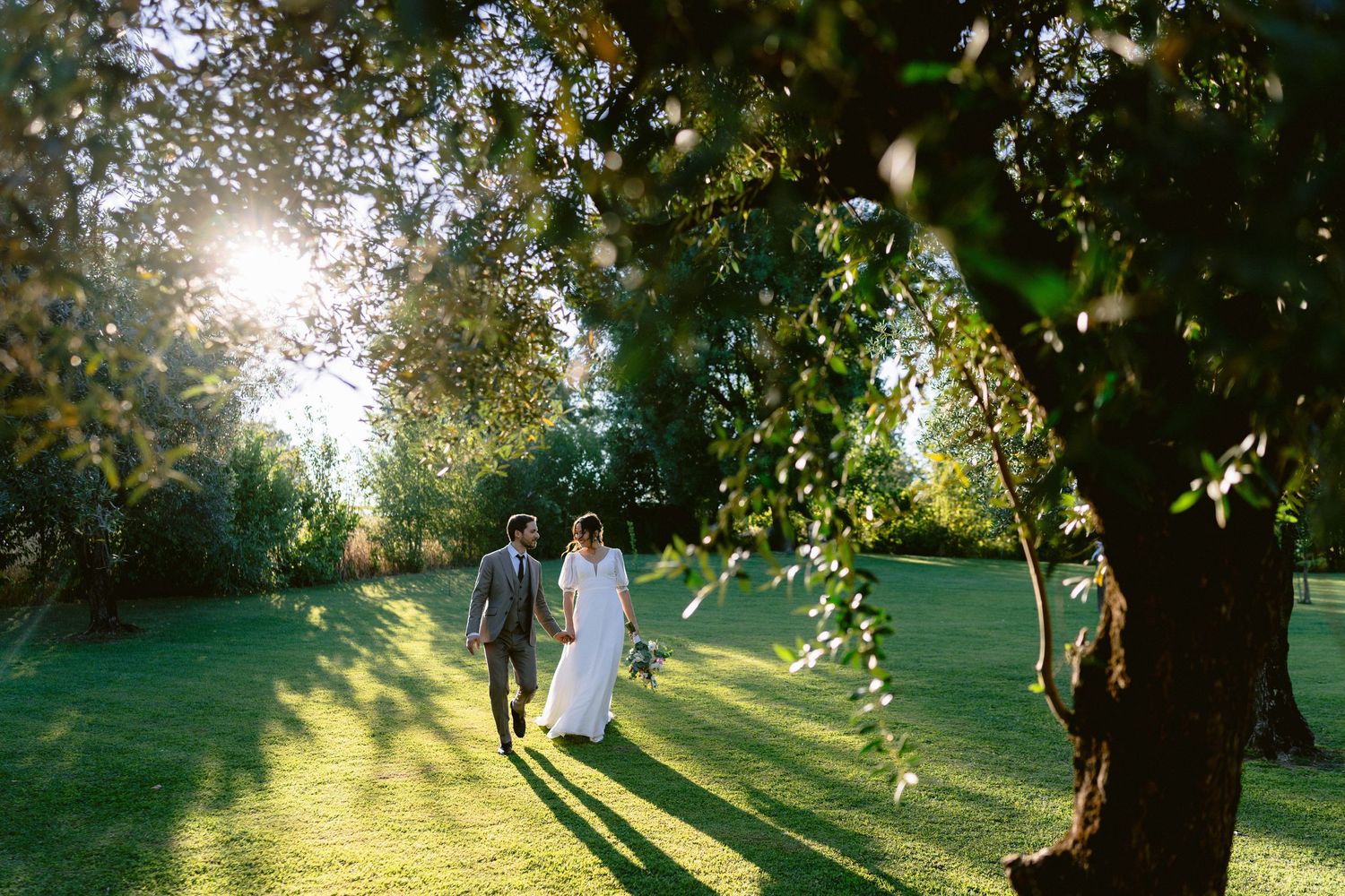 Golden sunlight streams through trees as a couple walks in a garden setting.