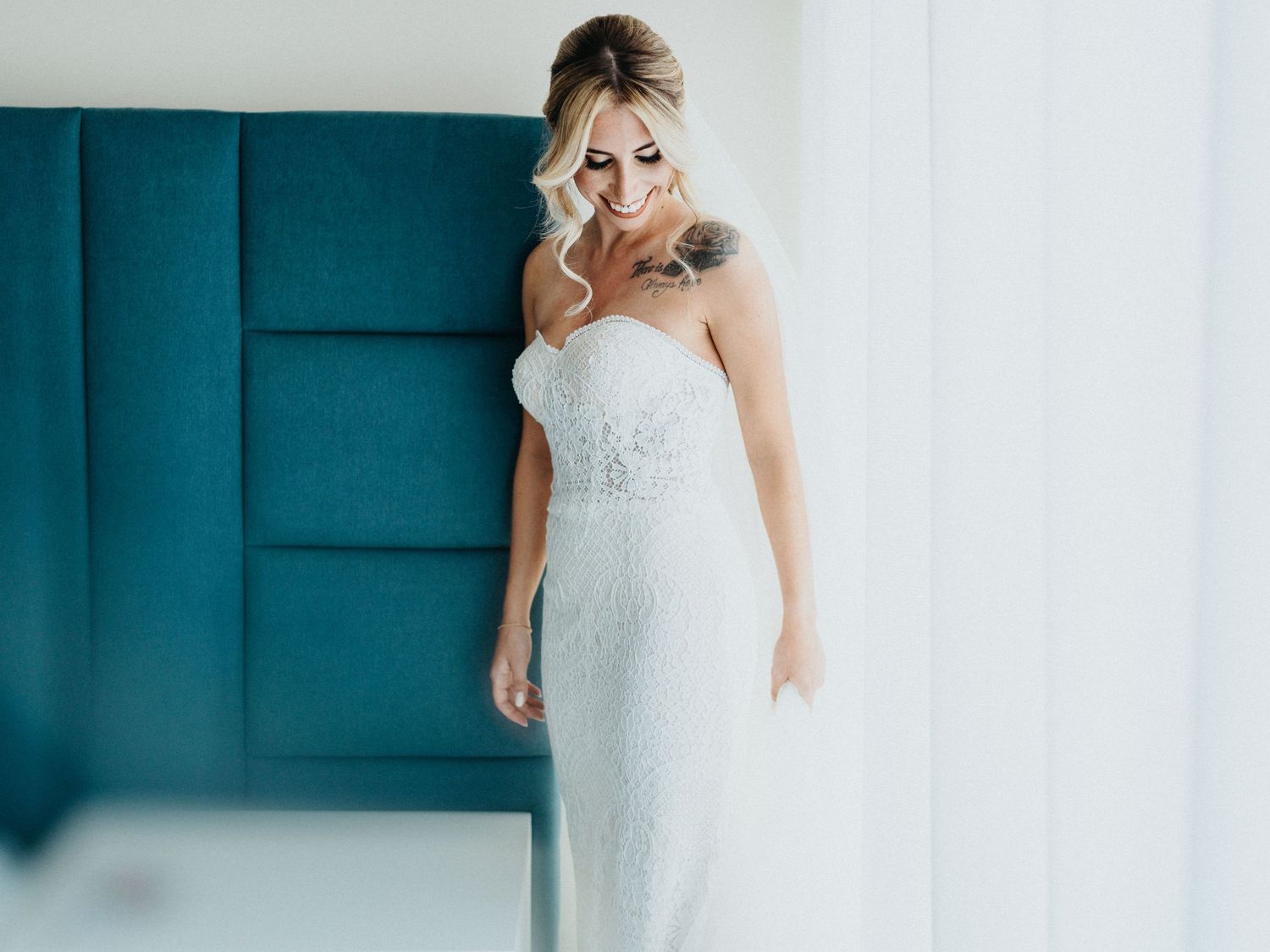 Elegant bride in white lace gown poses against turquoise wall in romantic bridal portrait series.