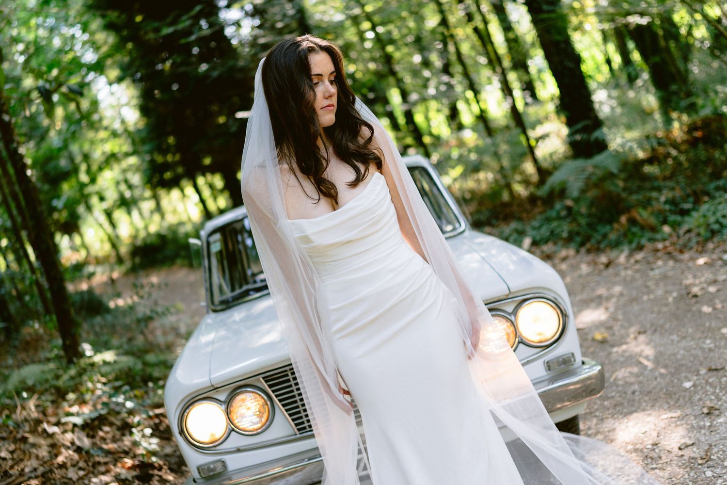A bride in a white dress poses next to a vintage car in a sunlit forest setting.