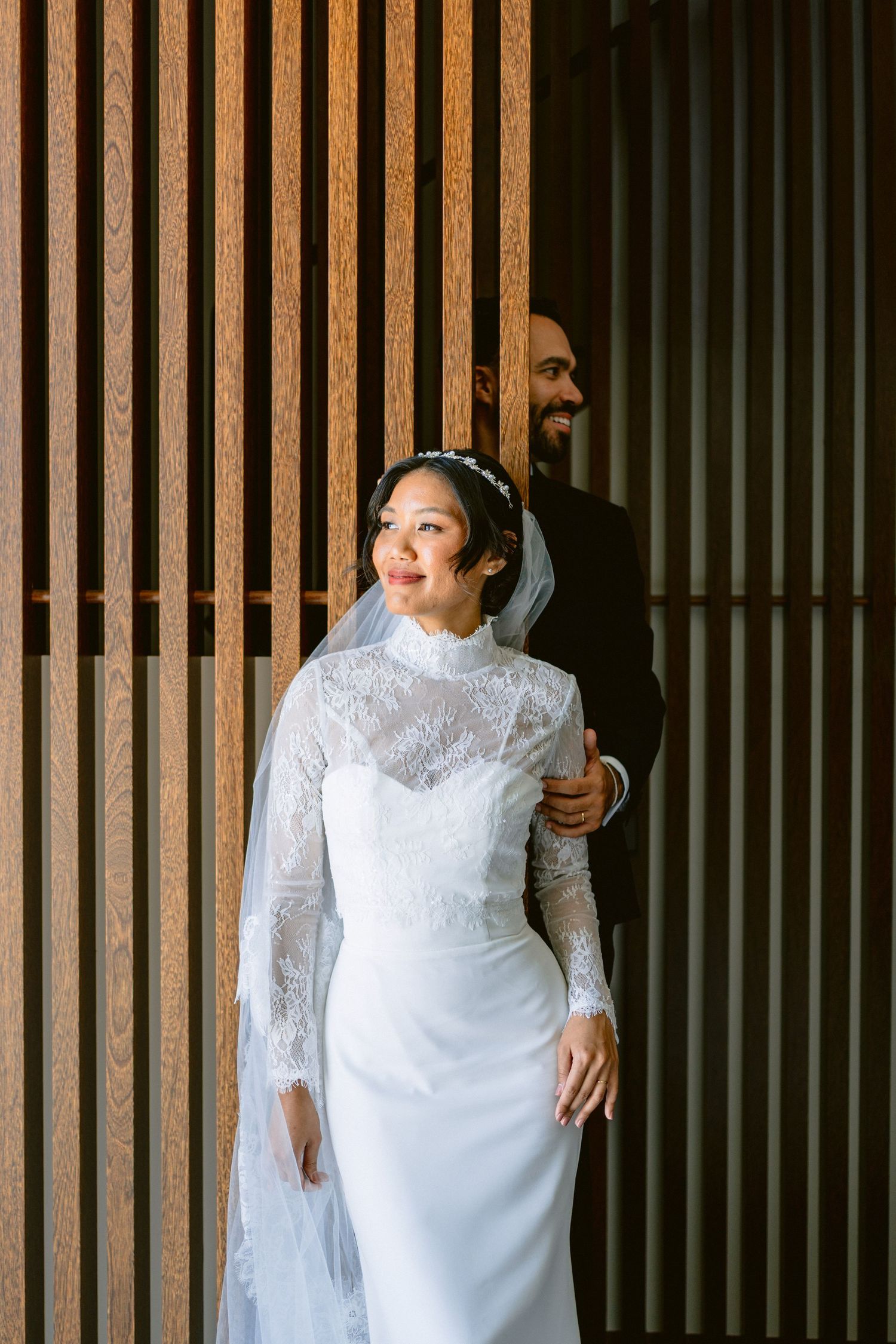 Bride in lace gown poses against wooden slat wall in dramatic lighting.
