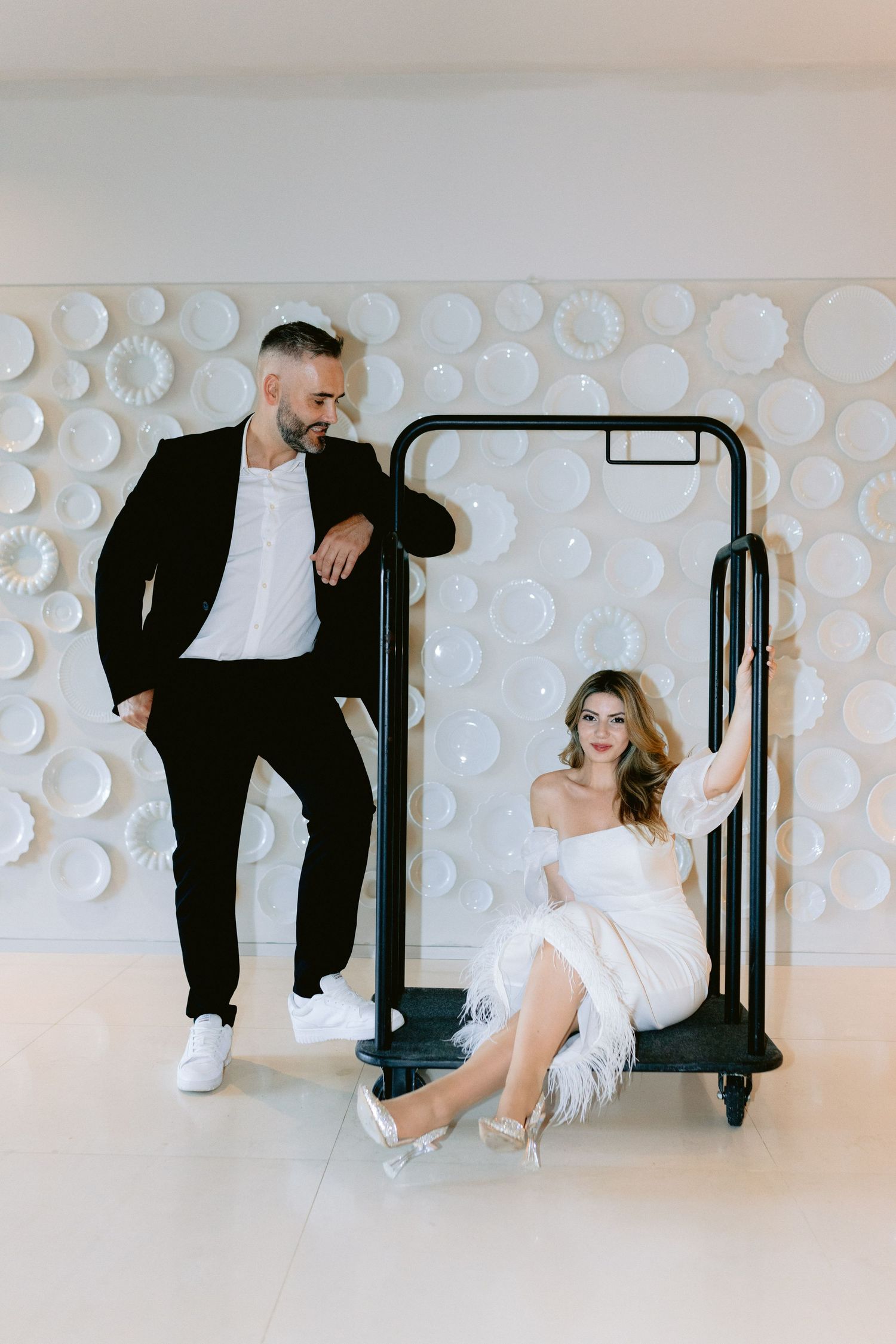 A stylish couple poses with a luggage cart against a white textured wall.
