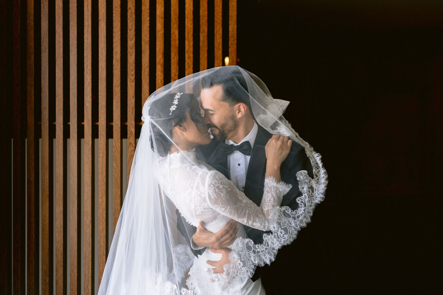 Dramatic silhouette of bride with flowing veil against wooden slatted wall.