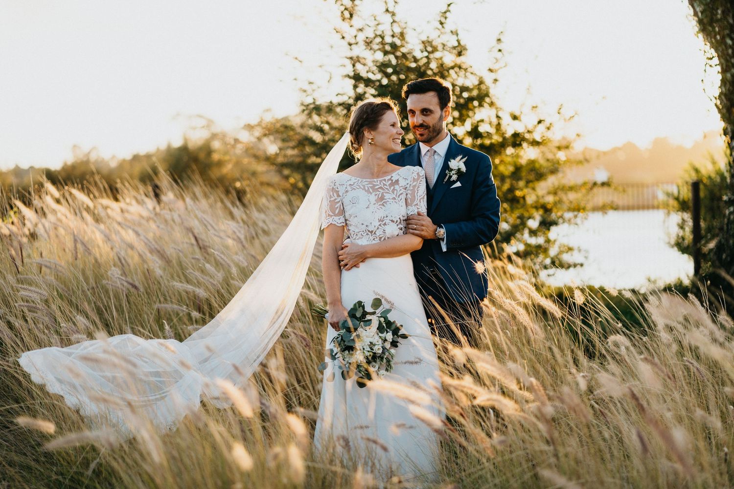 Couple in wedding attire walking through golden grass at sunset.