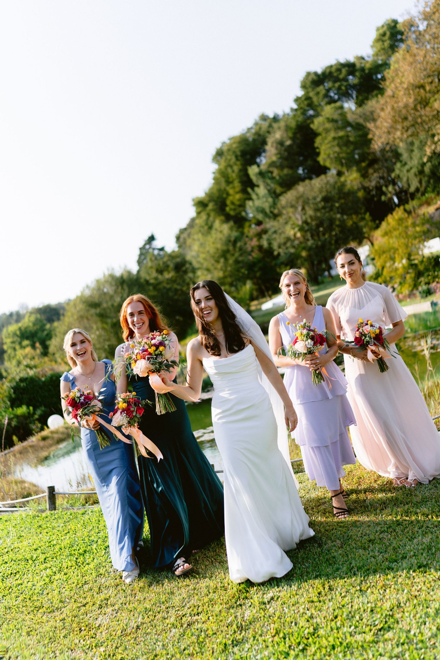 Wedding party in mixed blue and white attire poses playfully on grassy riverbank.