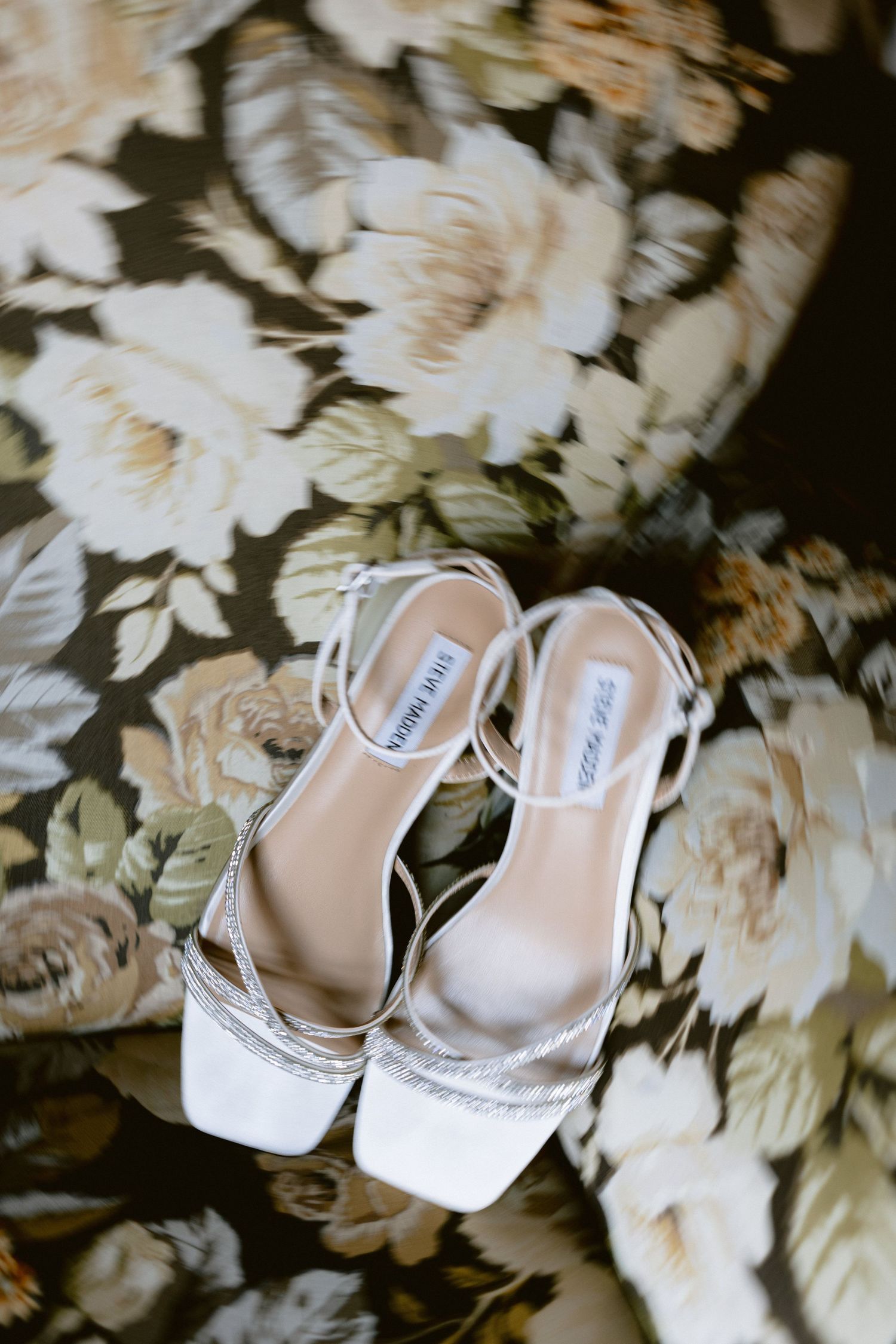 White bridal shoes with delicate straps rest on a bed of white flower petals.