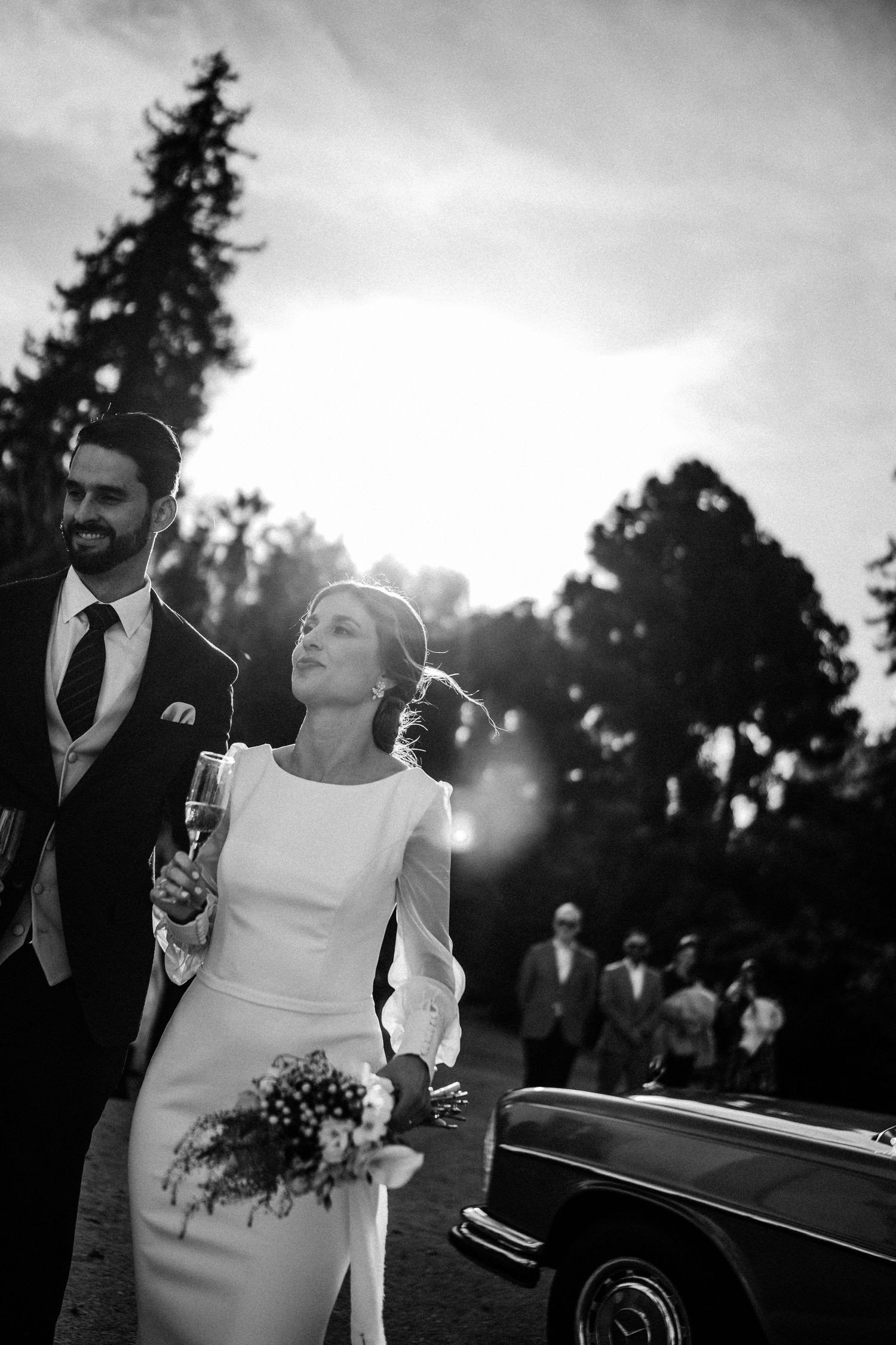 A black and white wedding portrait with a classic car and forest backdrop.