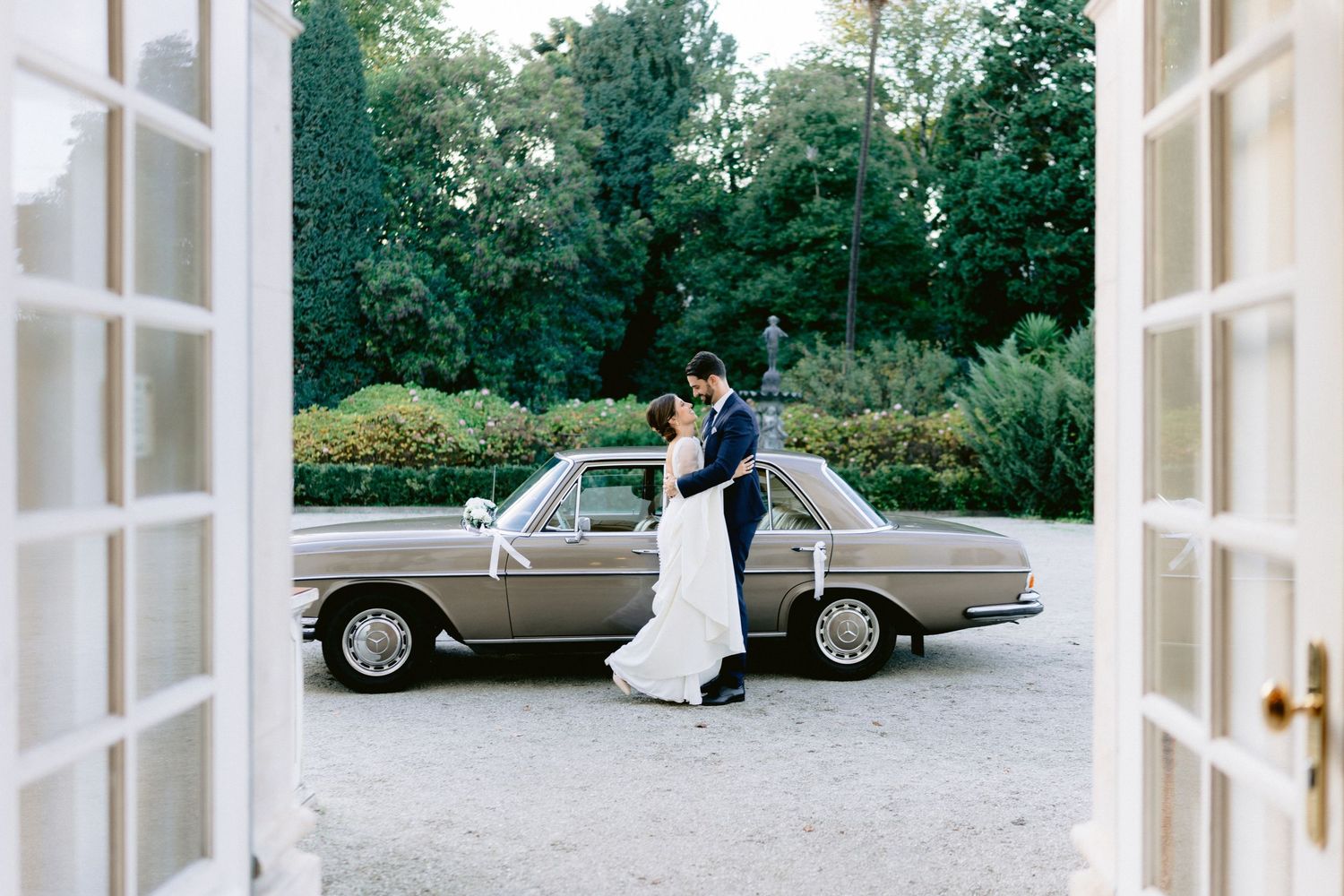 Elegant wedding couple poses near classic car and white French doors in outdoor garden setting.