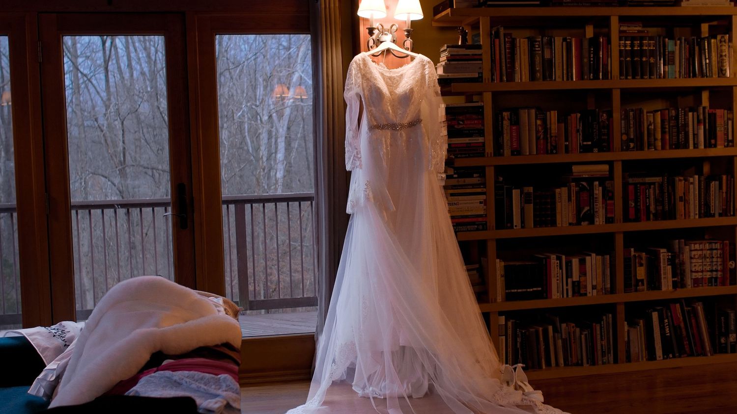 An elegant long-sleeved white wedding gown displayed on a hanger in a cozy room with bookshelves.