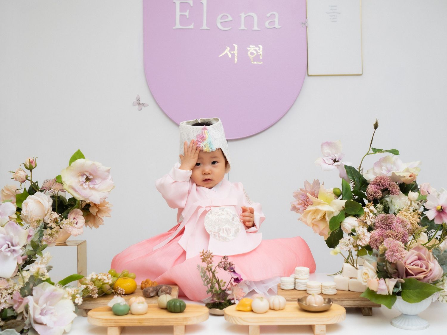 Baby girl in pink hanbok at dol (first birthday) celebration with flowers and traditional Korean doljabi items.