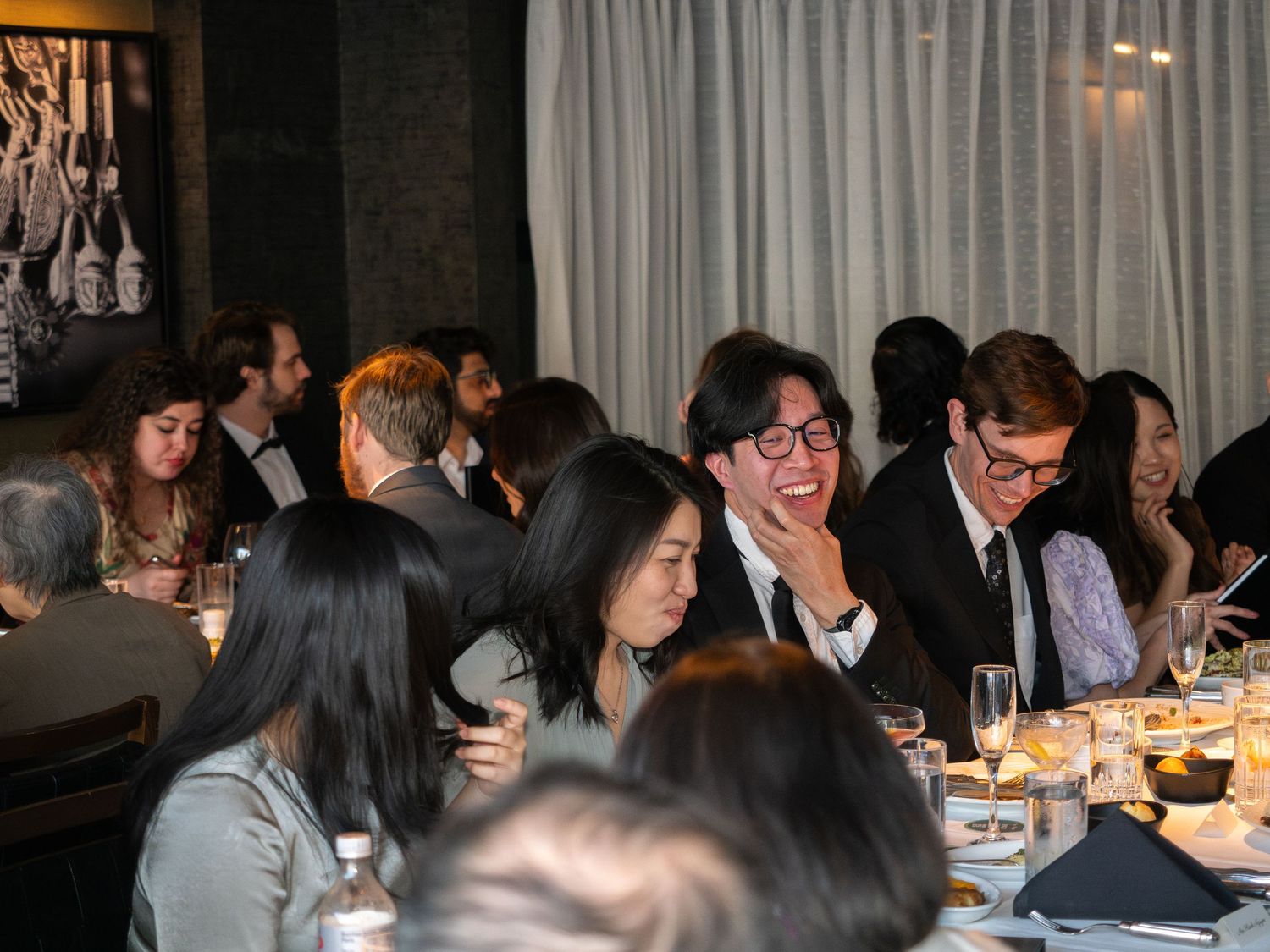 Guests laughing and socializing at an elegant dinner party with candlelit tables and formal attire.