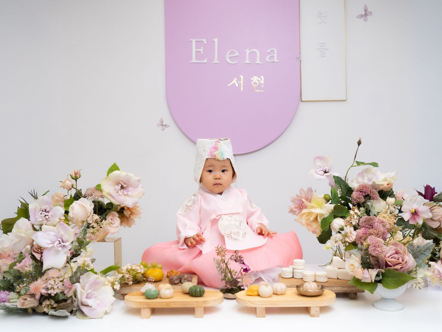 Baby girl Elena sits in traditional Korean hanbok dress surrounded by flowers at her doljabi first birthday celebration.