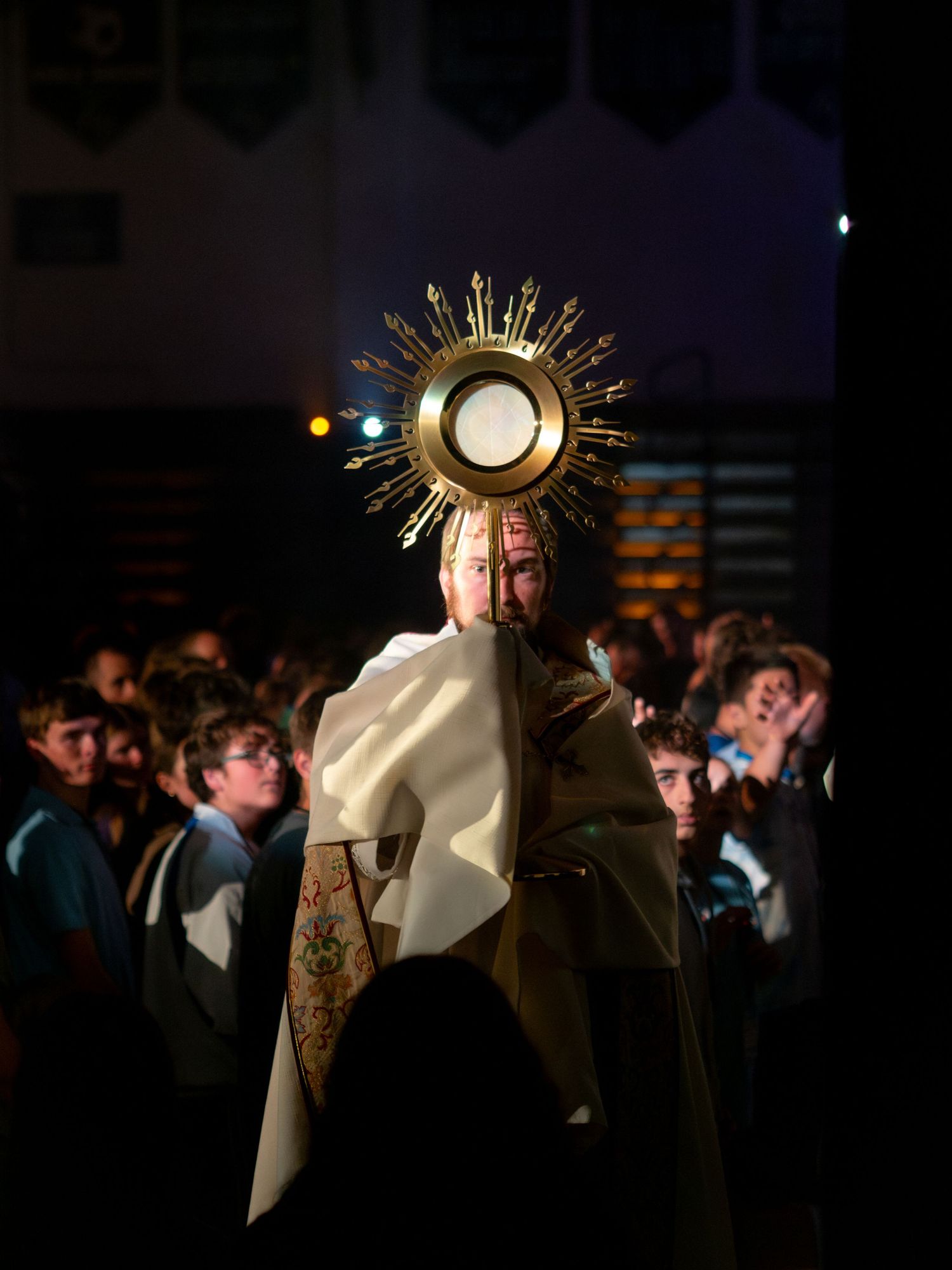 Catholic priest carrying ornate gold monstrance during Eucharistic procession before large crowd.