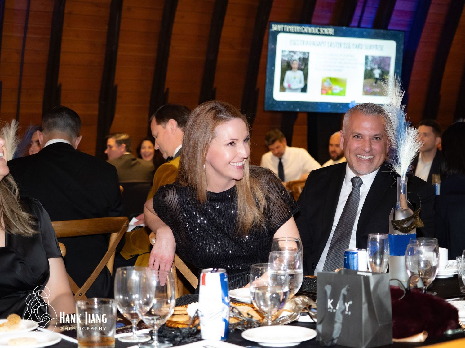 Elegantly dressed couple smiling at a formal gala dinner event with table settings and presentation screen in background.