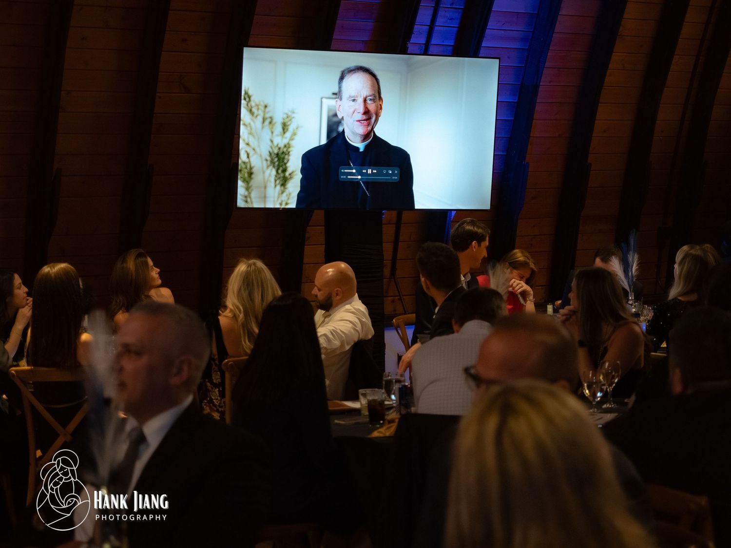 Guests attend a formal gala event watching a speaker on a large video screen in an elegant brick venue.