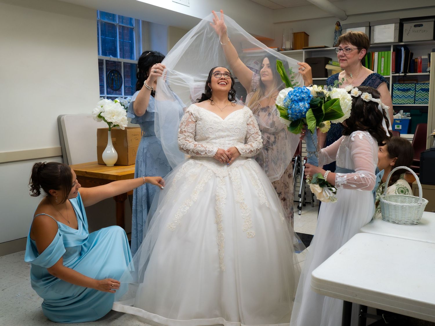 Bride in white lace gown smiles as bridesmaids carefully arrange her veil before the ceremony.
