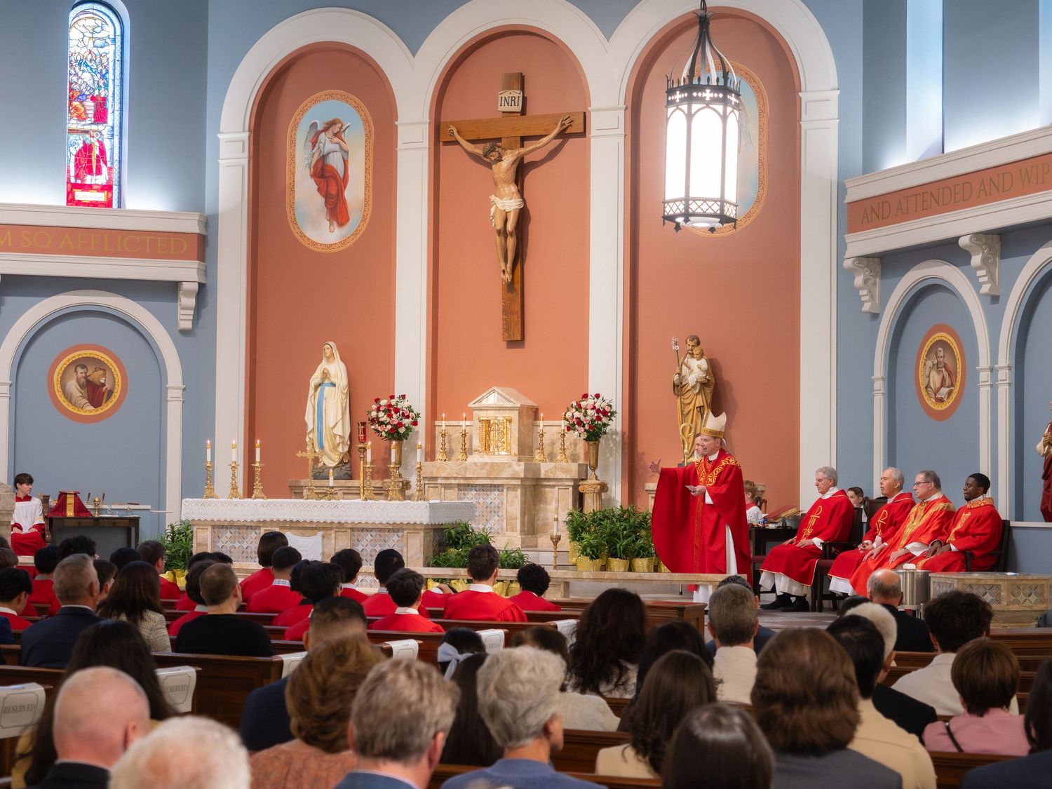 Catholic Mass ceremony with clergy in red vestments at ornate altar with crucifix and floral arrangements.