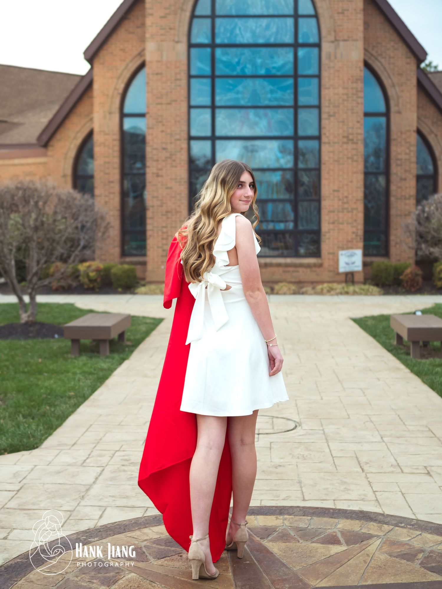 Young woman in white dress with red graduation stole poses in front of brick church building for senior photos.