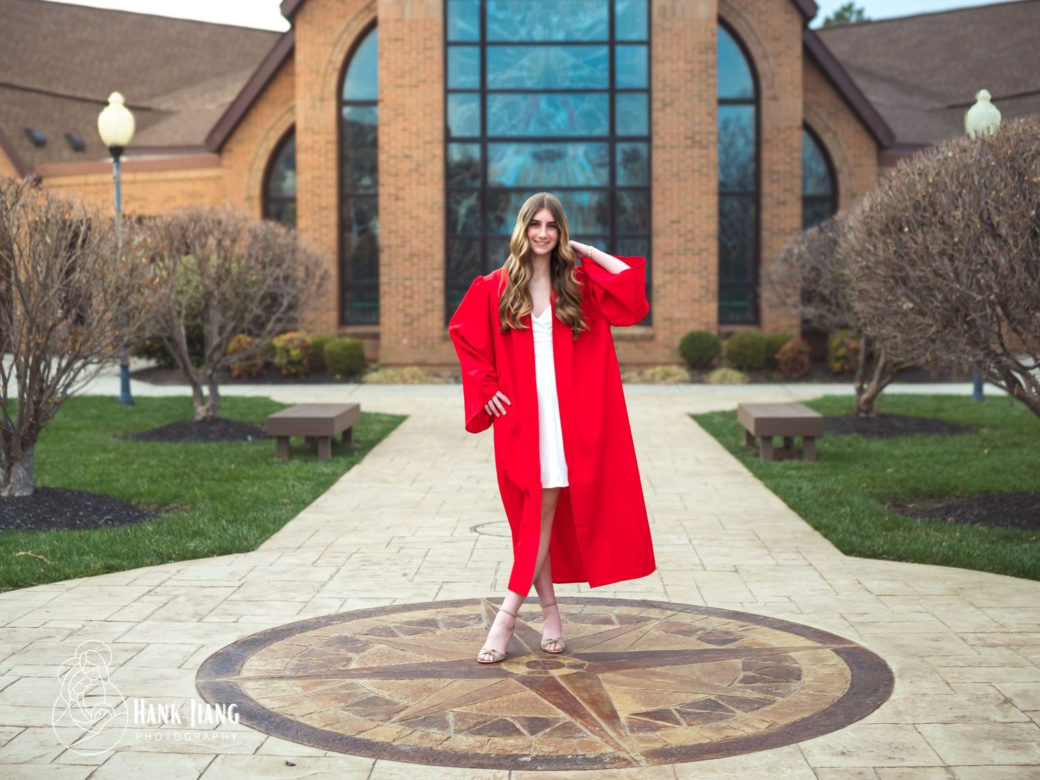 Young woman in red graduation gown and white dress posing on decorative pavement outside brick building.