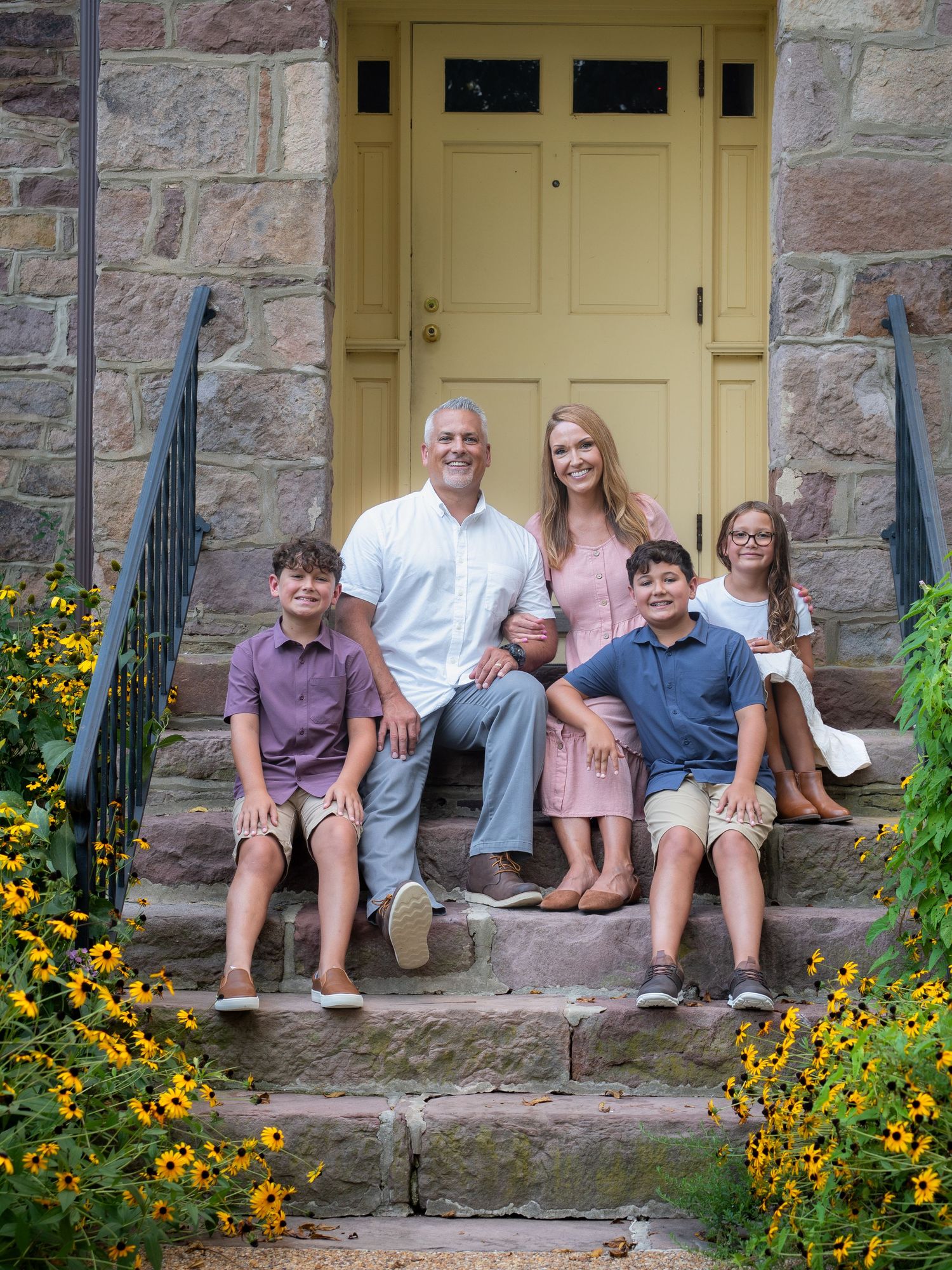 Happy family of five sitting on stone steps by a yellow door surrounded by blooming yellow flowers.