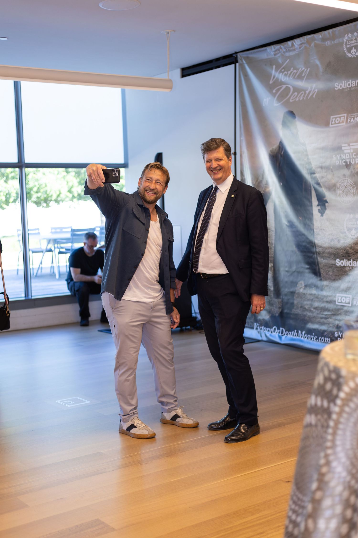 Two men smiling and taking a selfie indoors near a movie poster backdrop at a casual event.