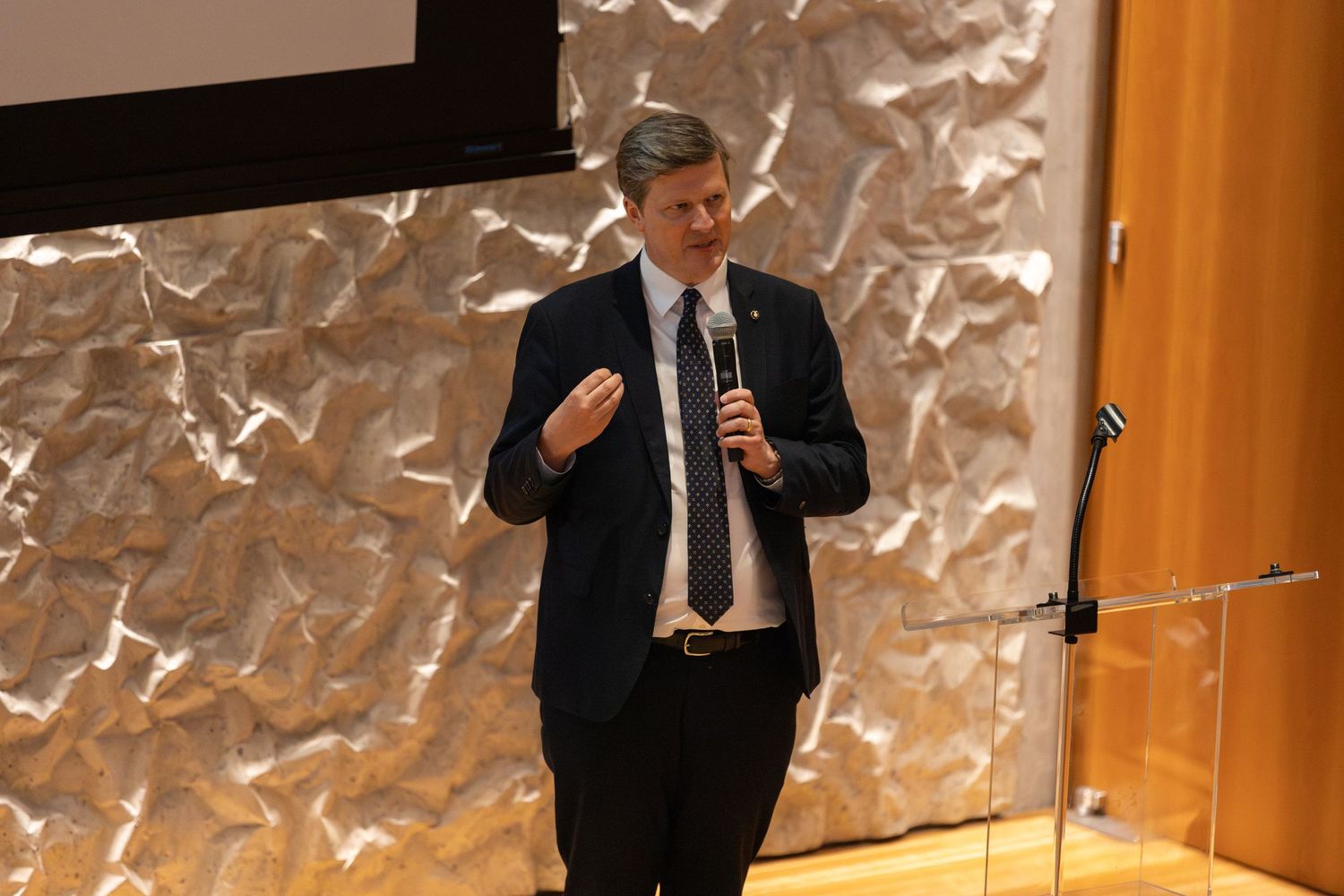 Professional speaker in navy suit holding microphone presenting at corporate event with textured stone wall backdrop.