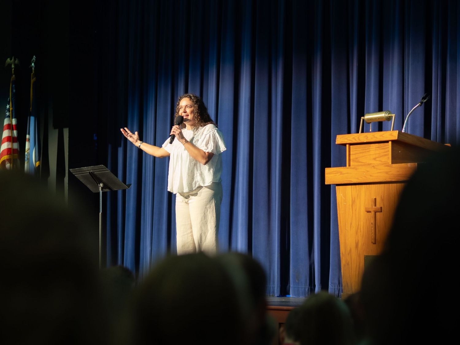 Woman in white outfit speaking at podium on stage with blue curtain backdrop at religious event.