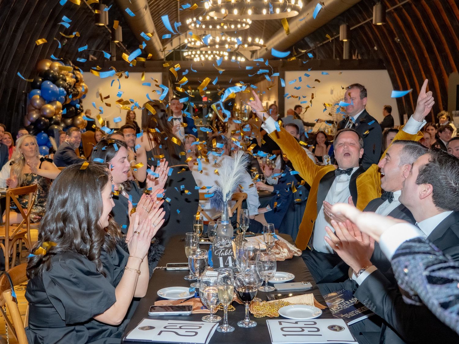 Guests cheer and applaud as colorful confetti rains down during a festive indoor celebration dinner.
