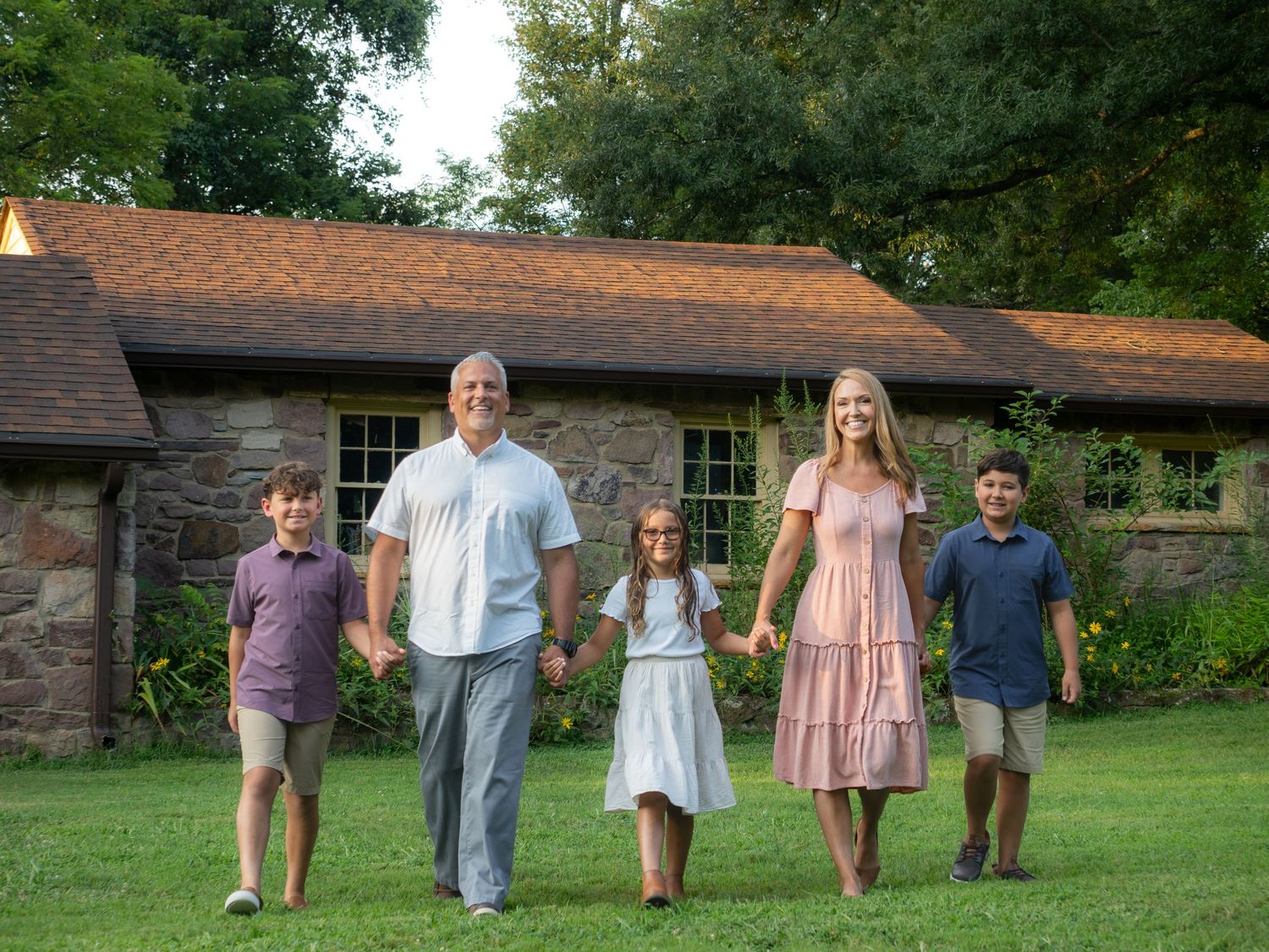 Family of five holding hands walking on green lawn in front of stone building.