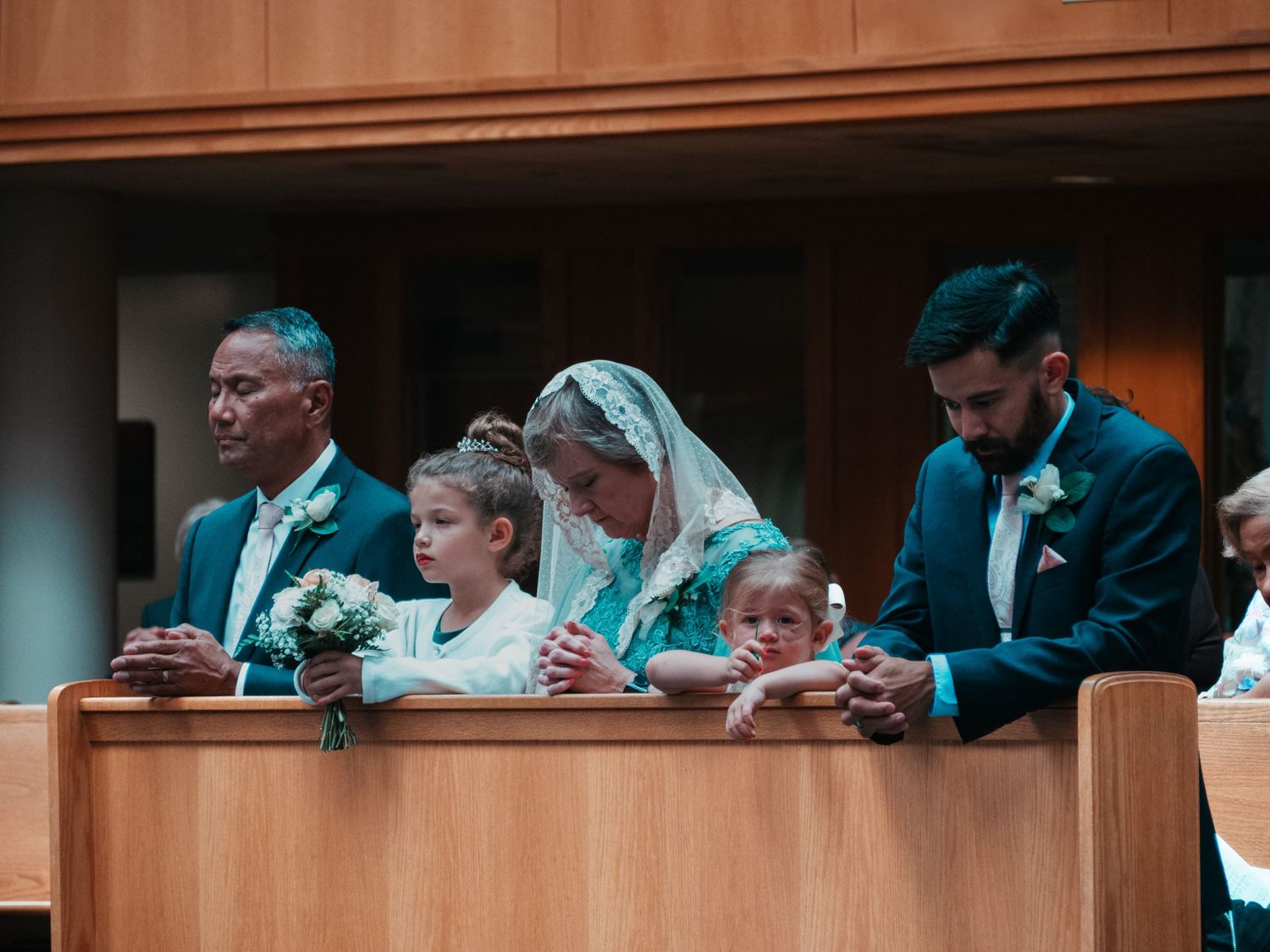 Wedding guests kneel in prayer at church pews during a religious ceremony, with bride in veil and children present.