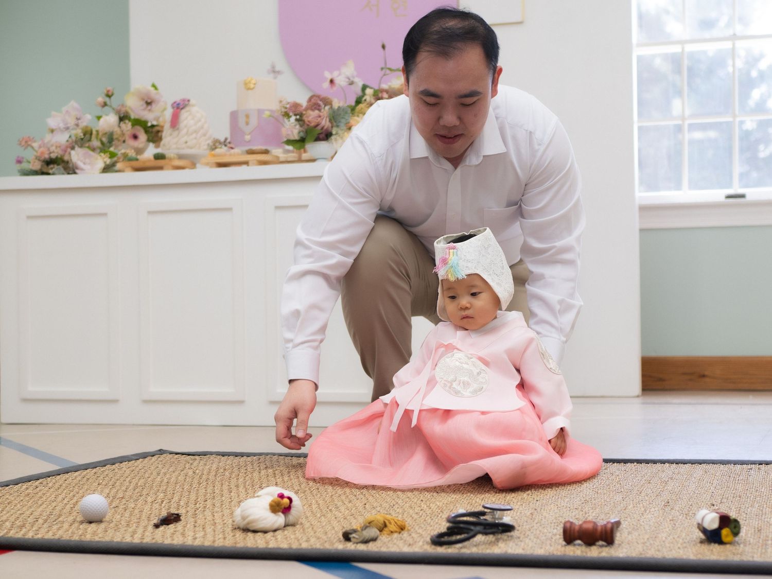 Father helping baby girl in pink Korean hanbok during traditional Doljabi ceremony with items spread on floor mat.