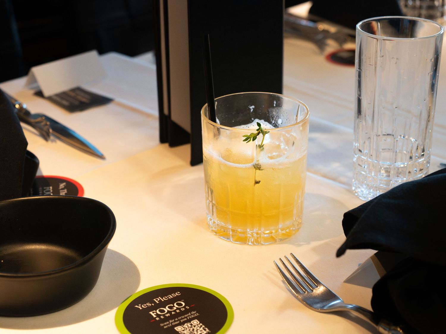 A golden cocktail garnished with herbs in a rocks glass on a white bar counter with utensils and a menu card.