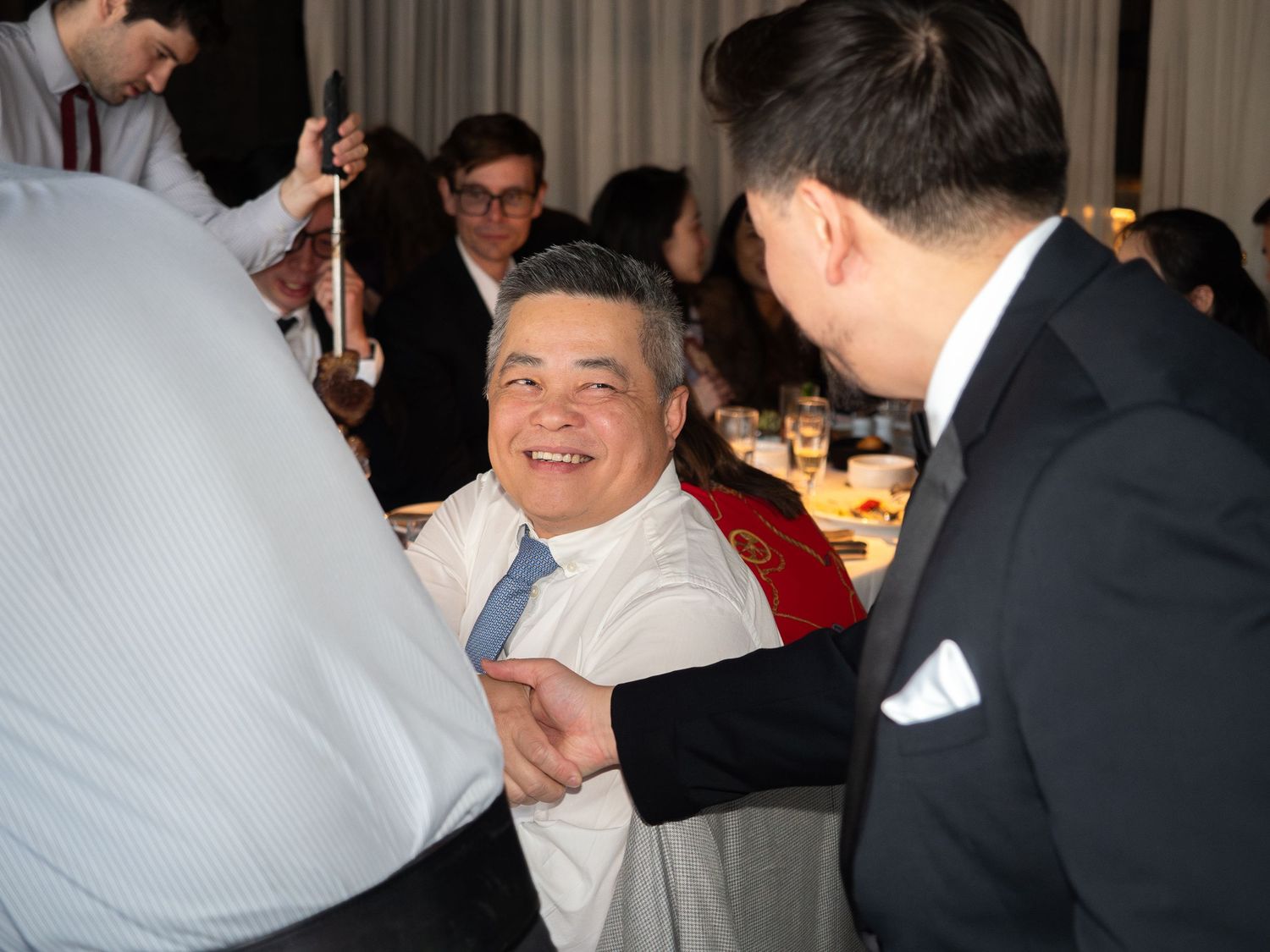 Smiling man in white shirt and blue tie seated at a formal dinner event surrounded by guests in suits.