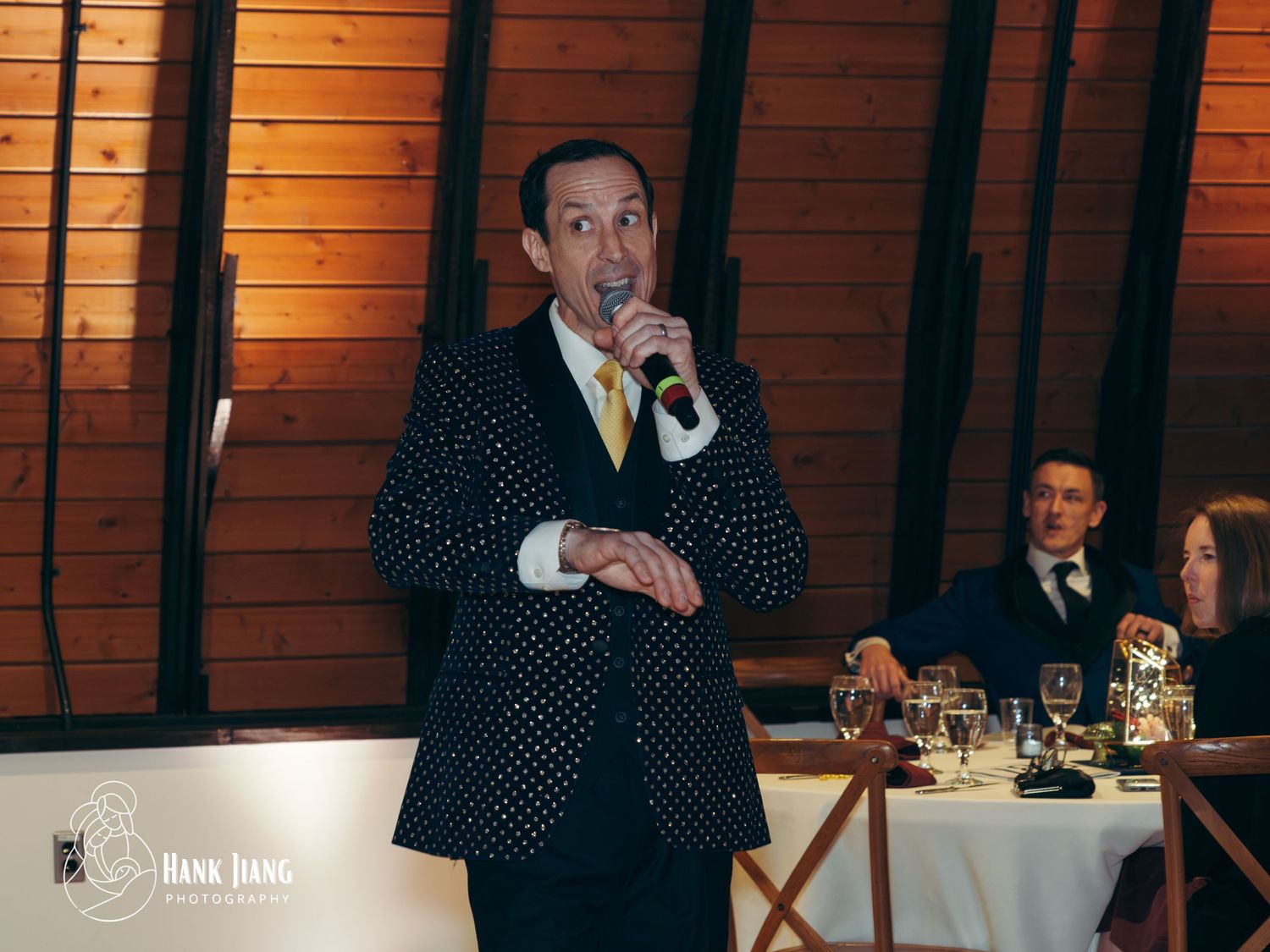 Man in polka dot suit speaking into microphone at elegant indoor event with guests seated at tables.