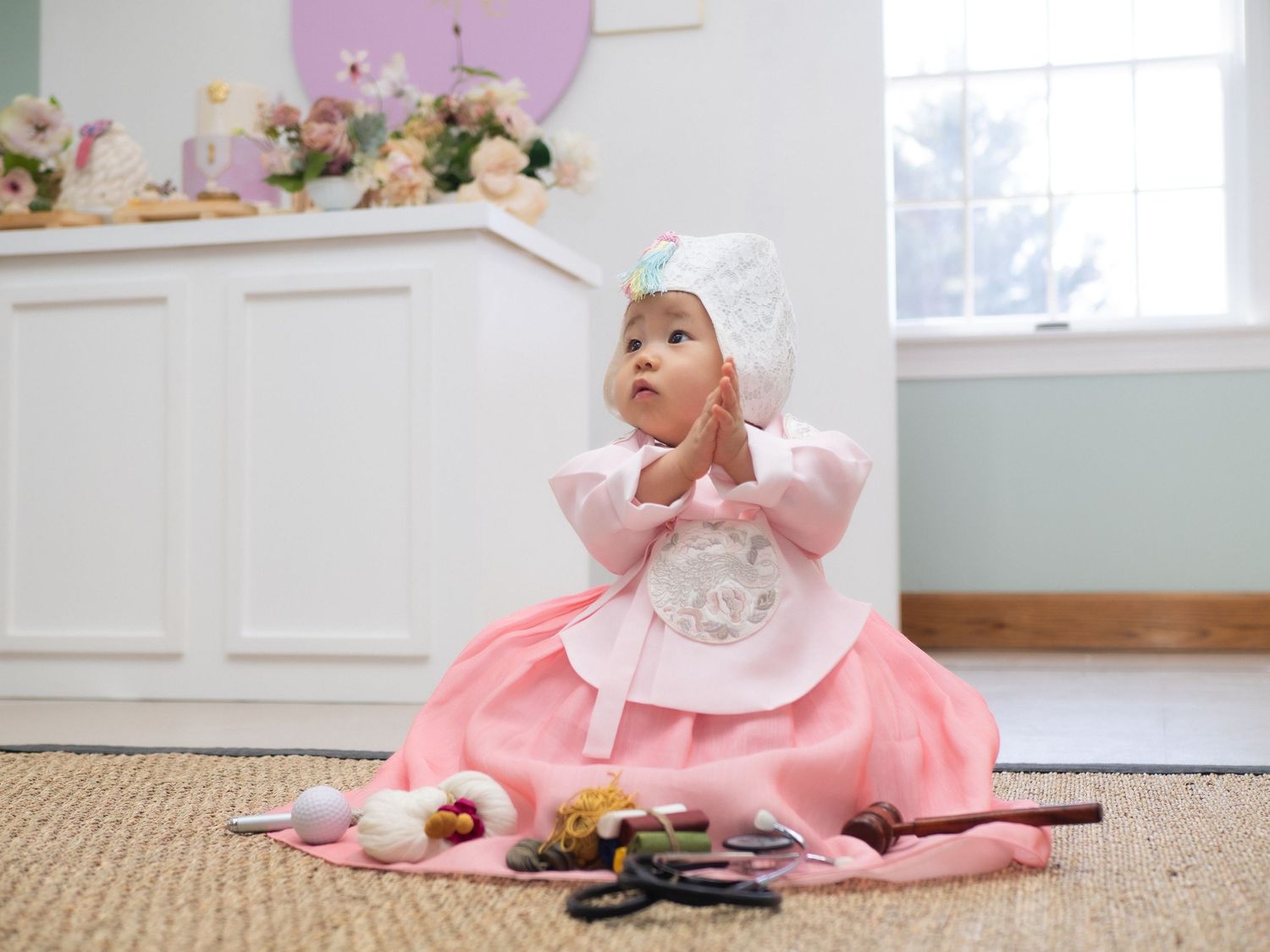 Baby girl in pink Korean hanbok dress and bonnet sitting on floor with traditional items during dol celebration.