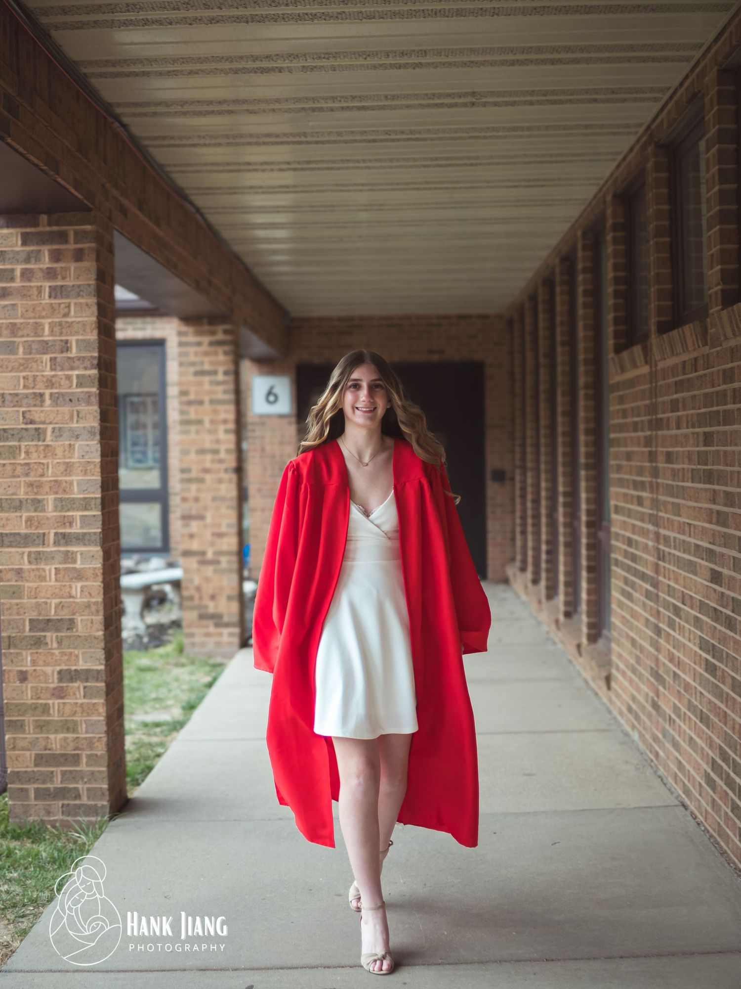 A smiling young woman walks in a red graduation gown over a white dress along a brick building corridor.