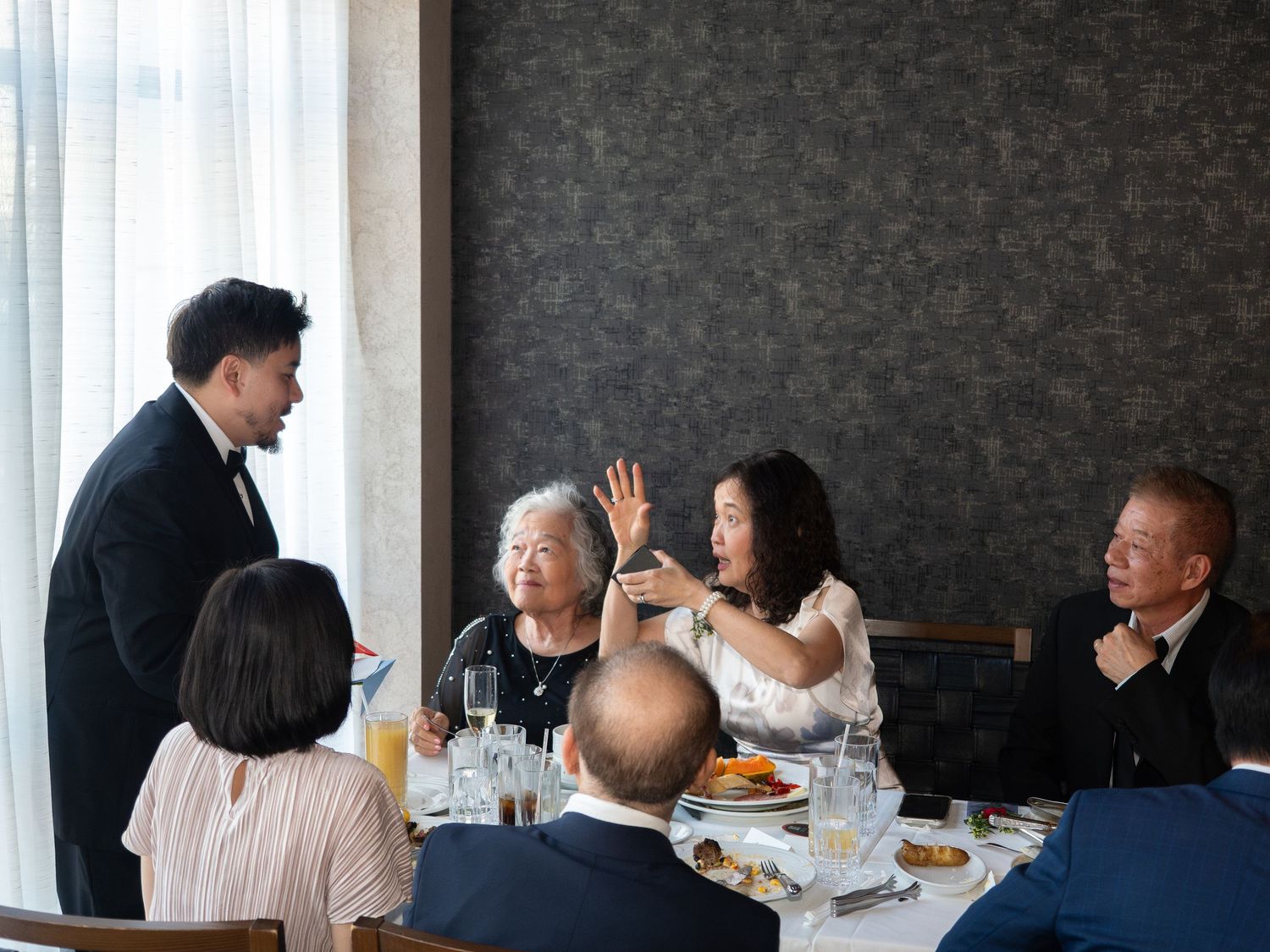 Group of Asian adults dining at a restaurant table, engaged in lively conversation with a suited waiter.