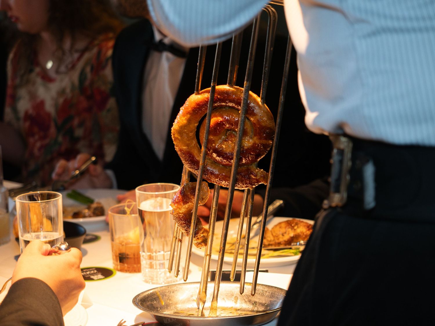 Waiter serving roasted meat on skewers over a metal bowl at an upscale restaurant dinner event.