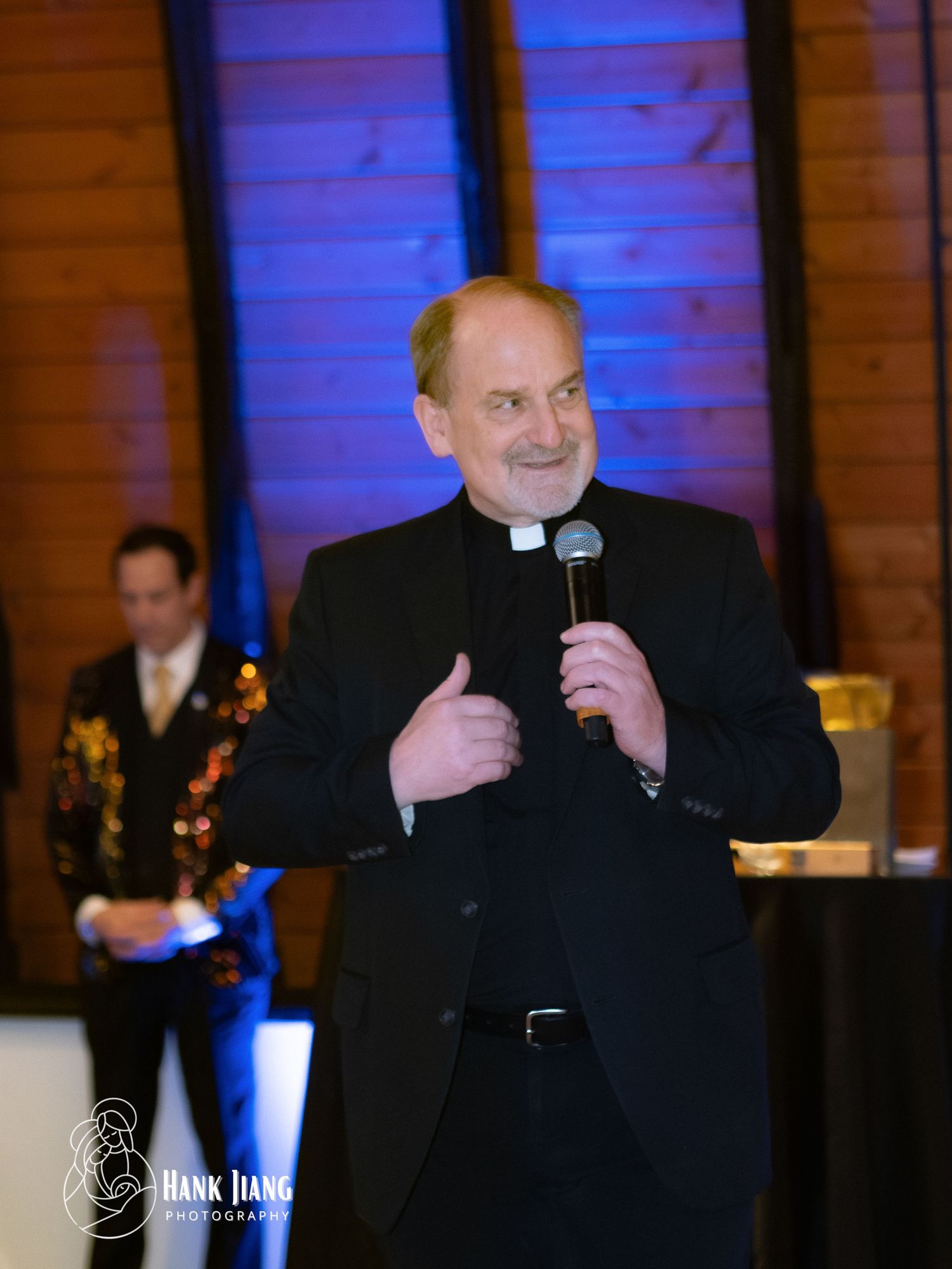 A smiling priest in black clergy attire holds a microphone and gives a thumbs up at an indoor event.