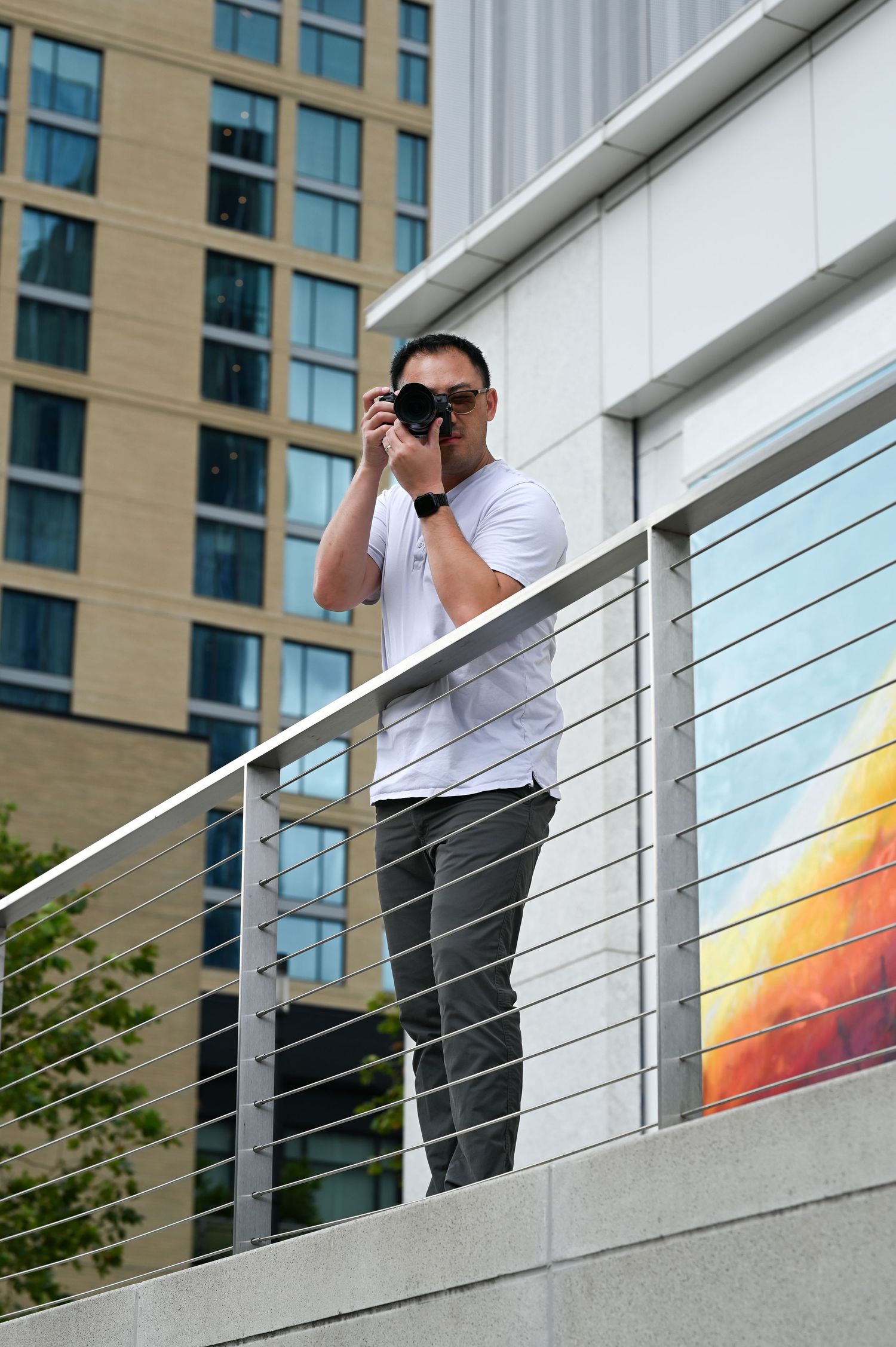 Hank Jiang, a male photographer, is in a white shirt capturing photos while standing on modern urban balcony with railing.