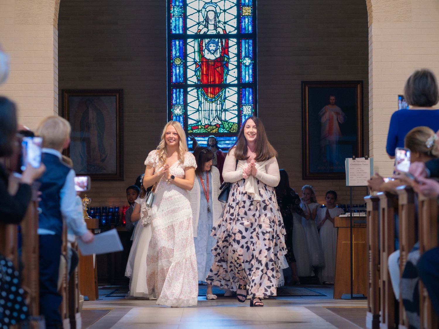 Two women walk down a church aisle during a wedding ceremony, with a stained glass window and guests in the background.