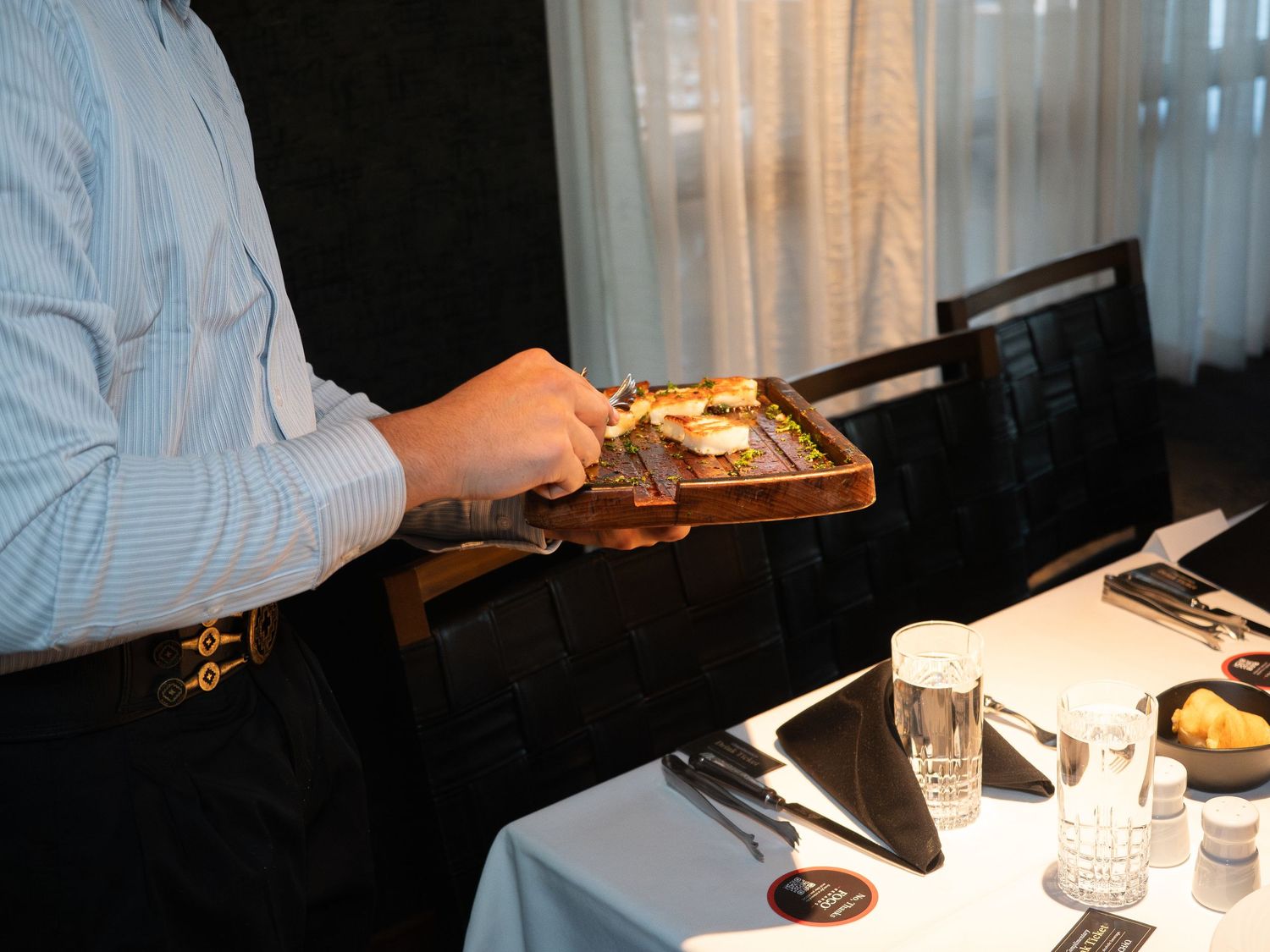 Waiter carrying a wooden tray with gourmet dish in an elegant restaurant setting with white tablecloths.