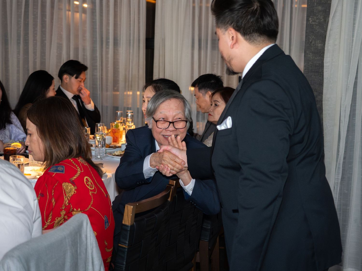 Elderly guest clapping hands at a formal dinner banquet, greeted by a man in a dark suit.
