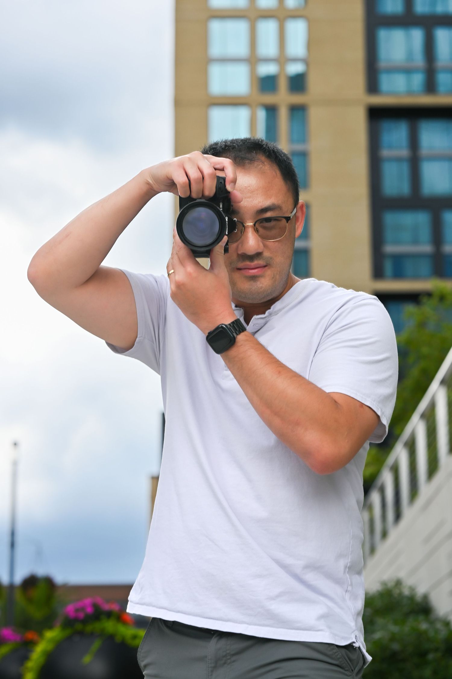 Hank Jiang, a male photographer, is in a white t-shirt holding a mirrorless camera outdoors with urban building background.