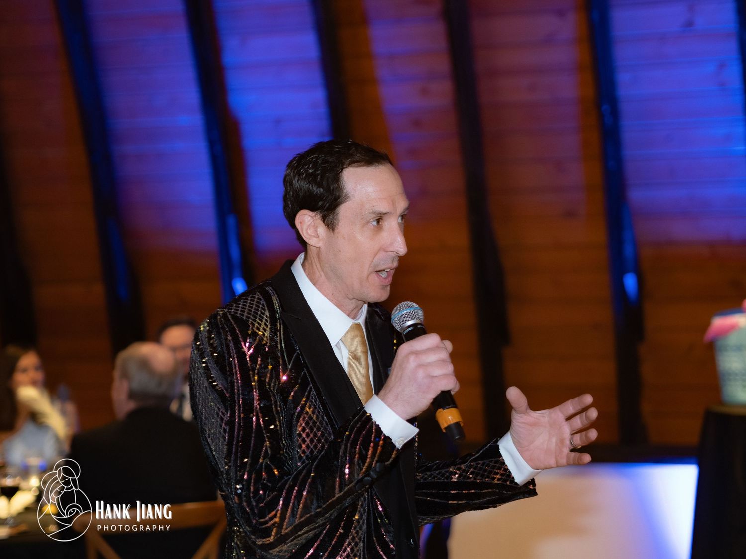 Man in sequined jacket speaking into microphone at formal event, gesturing expressively on stage.