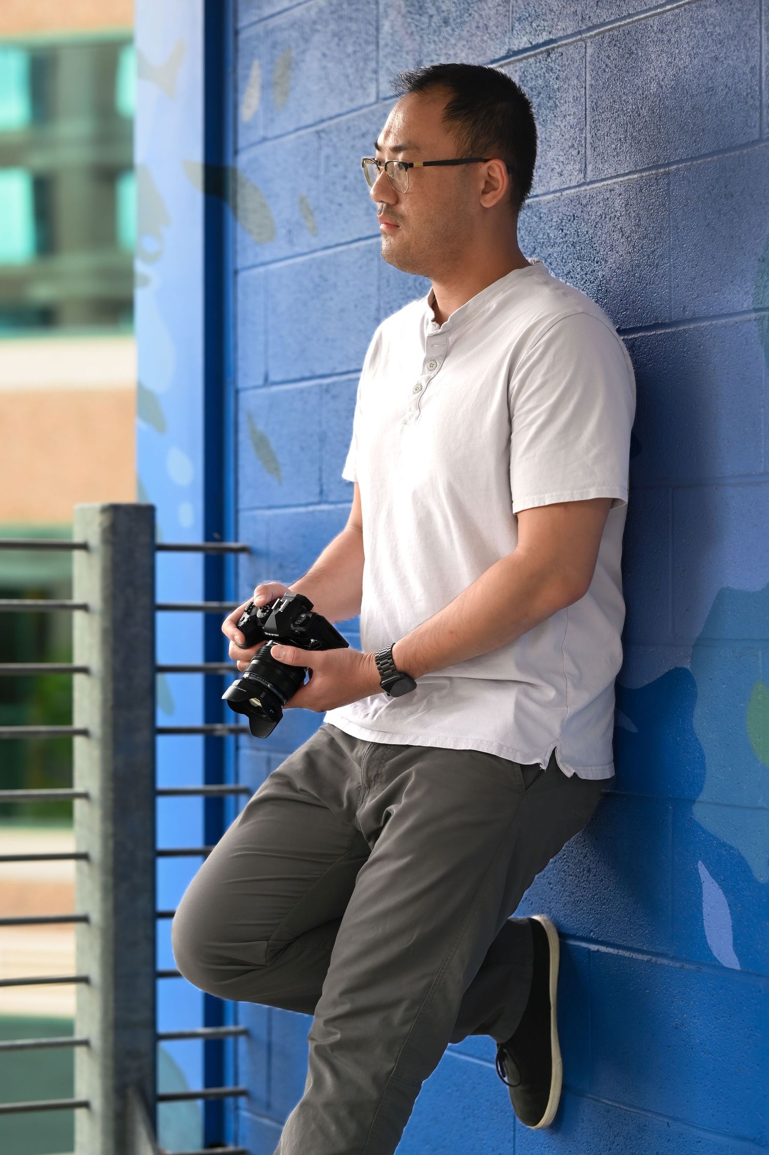 Hank Jiang, a male photographer, is leaning against blue wall holding camera in urban outdoor environment.