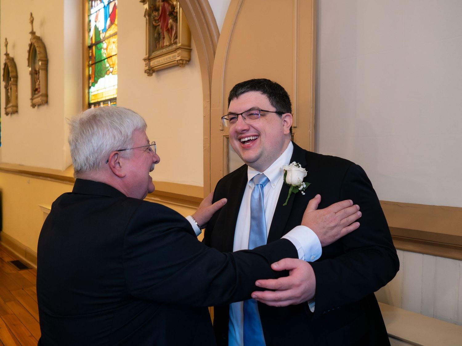 Groom laughing joyfully with an older gentleman inside a beautiful stained glass church.